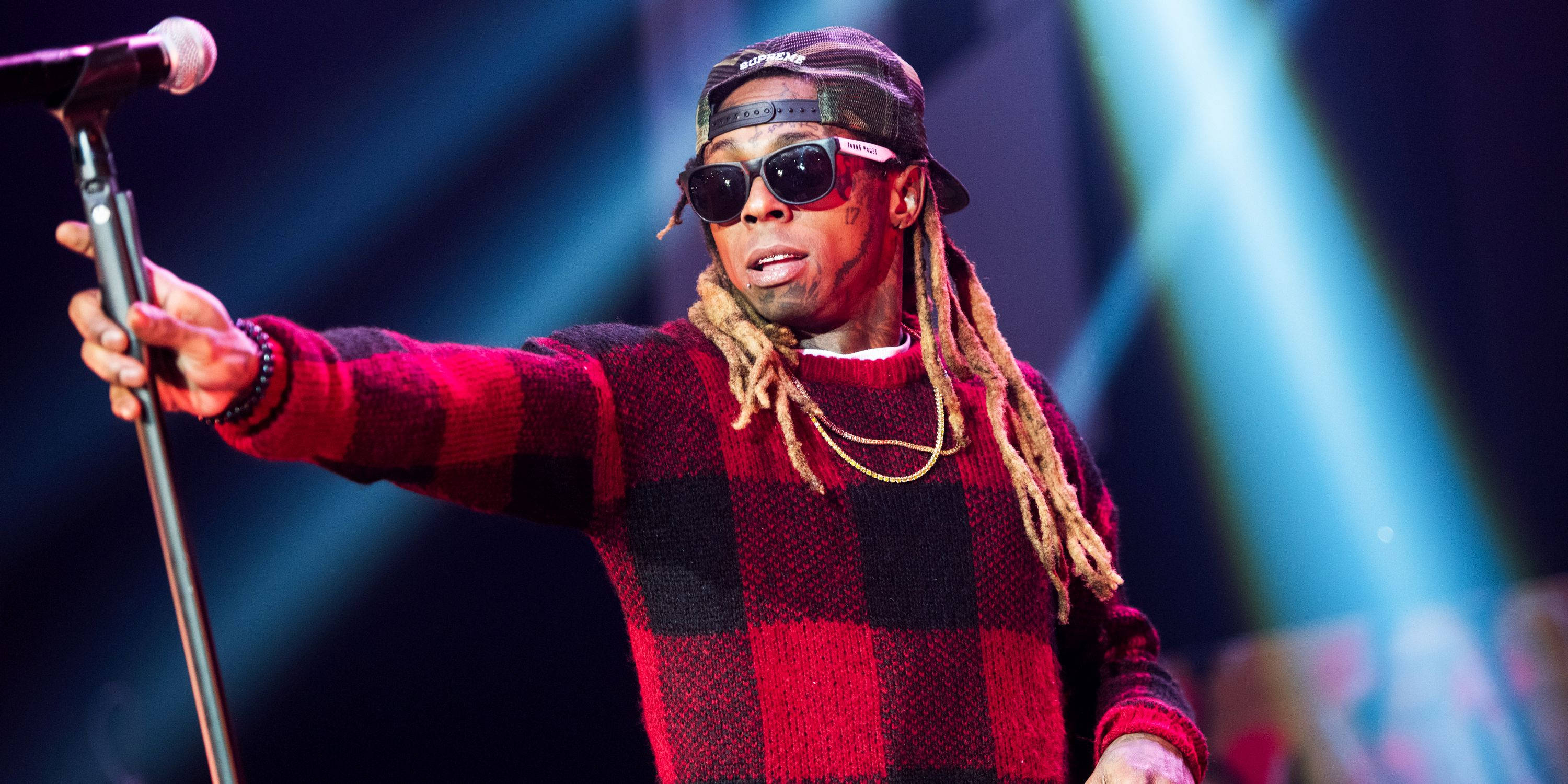 Lil Wayne 桌布 壁紙 — 一位穿著紅色格紋毛衣和太陽眼鏡的饒舌歌手在藍色聚光燈下拿著麥克風在舞台上表演。