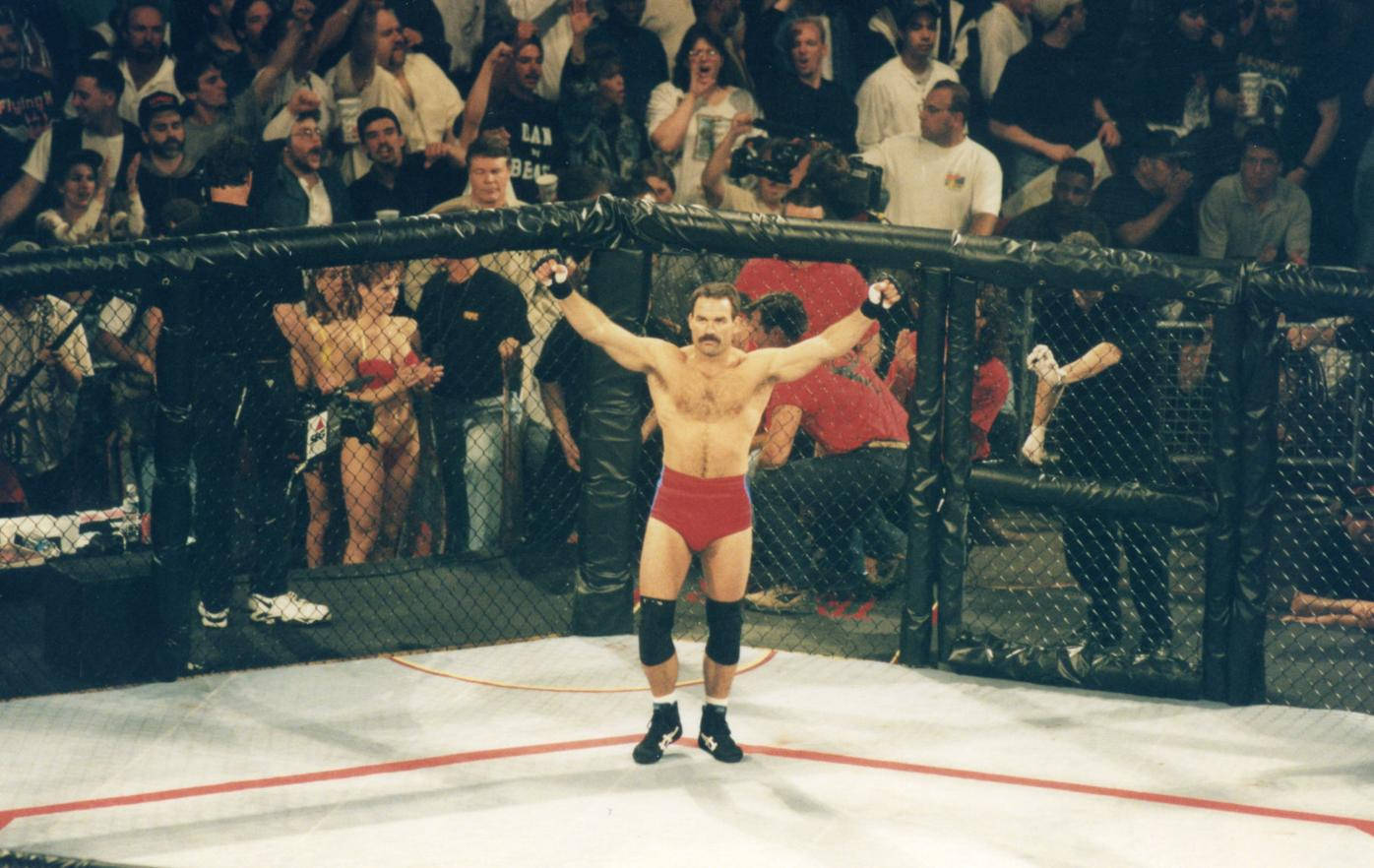 UFC 桌布 壁紙 — 一位穿著紅色短褲的摔角手在鐵籠中高舉雙臂慶祝勝利，興奮的觀眾在旁觀看。