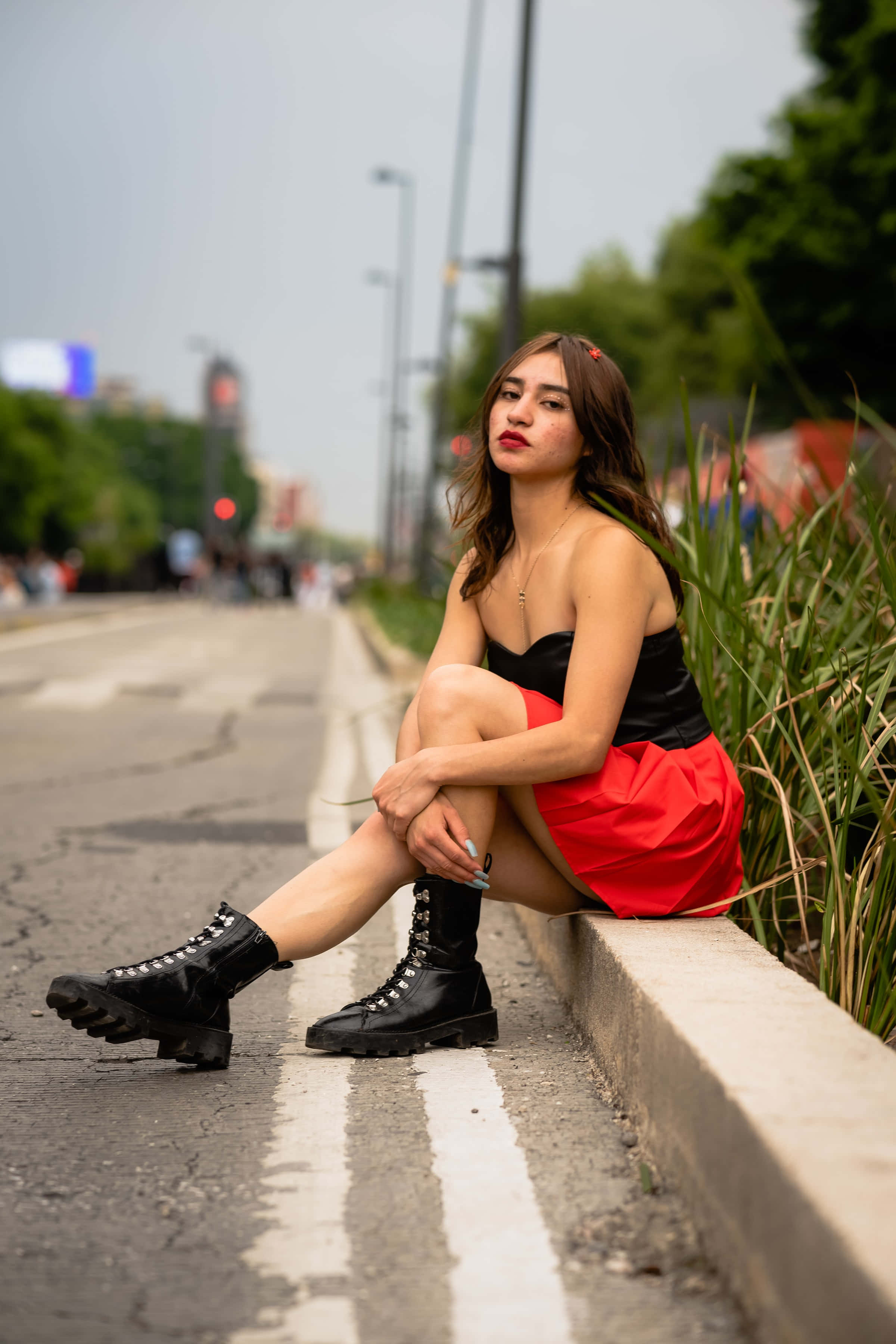 Woman Beautiful 桌布 壁紙 — 一位穿著紅色裙子和黑色靴子的年輕女性，坐在路邊石階上，若有所思，背景是模糊的城市景觀。