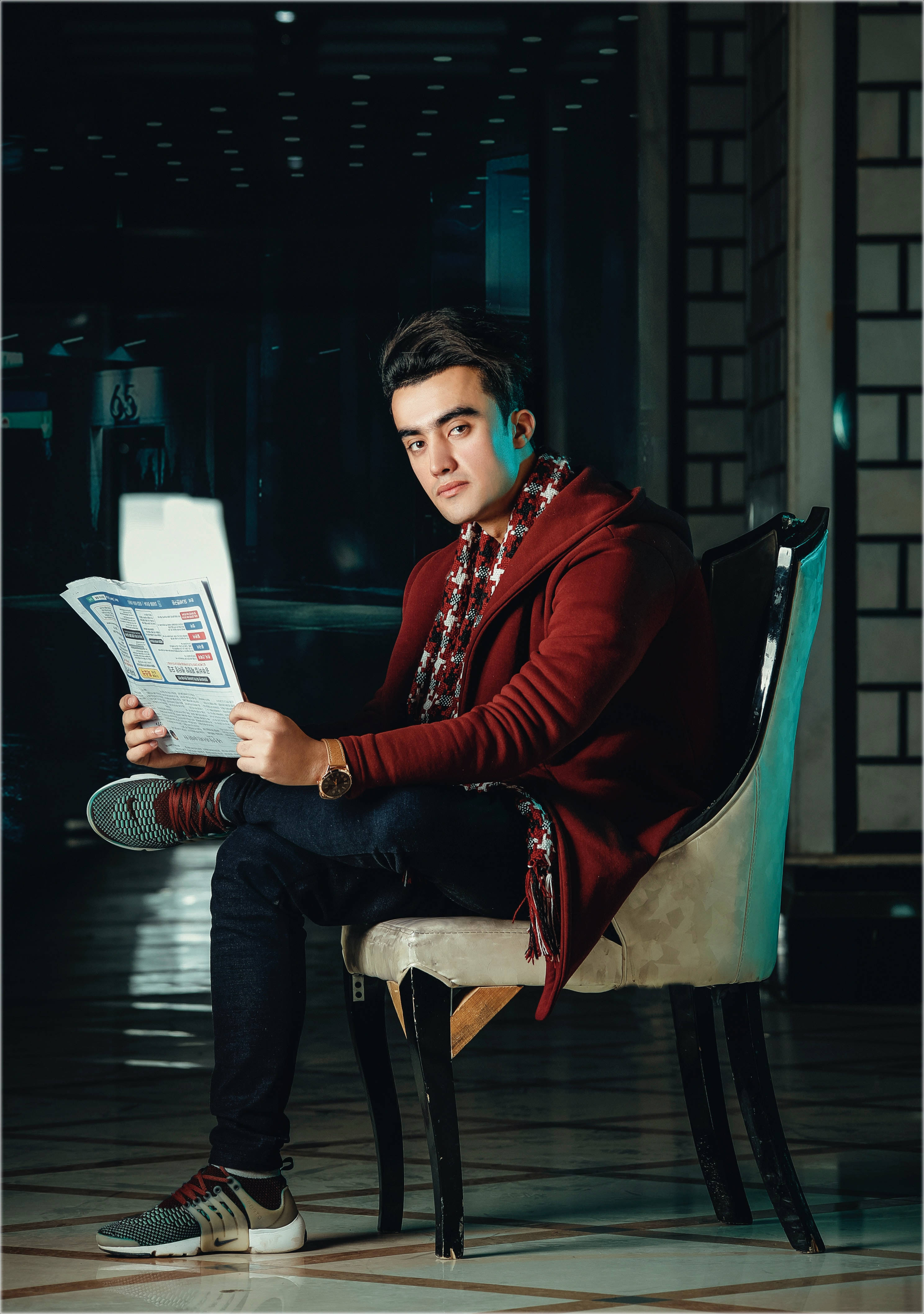 male model 桌布 壁紙 — 一位穿著紅色連帽衫的年輕男子坐在椅子上，在現代走廊中戲劇性的藍色和紅色燈光下閱讀報紙。
