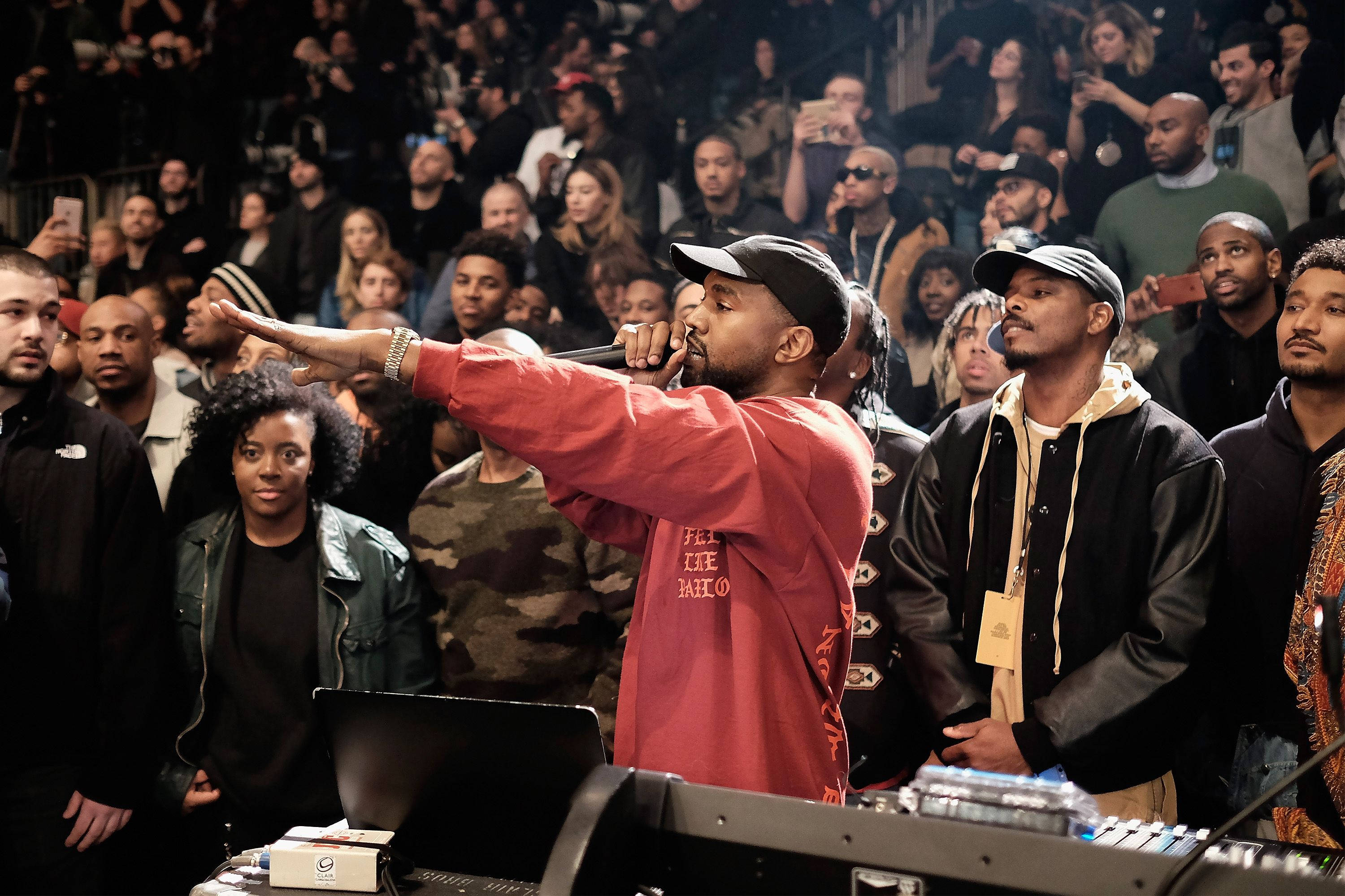 Kanye 桌布 壁紙 — 一位穿著紅色連帽衫的饒舌歌手在舞台上用麥克風表演，投入的觀眾專注地觀看。