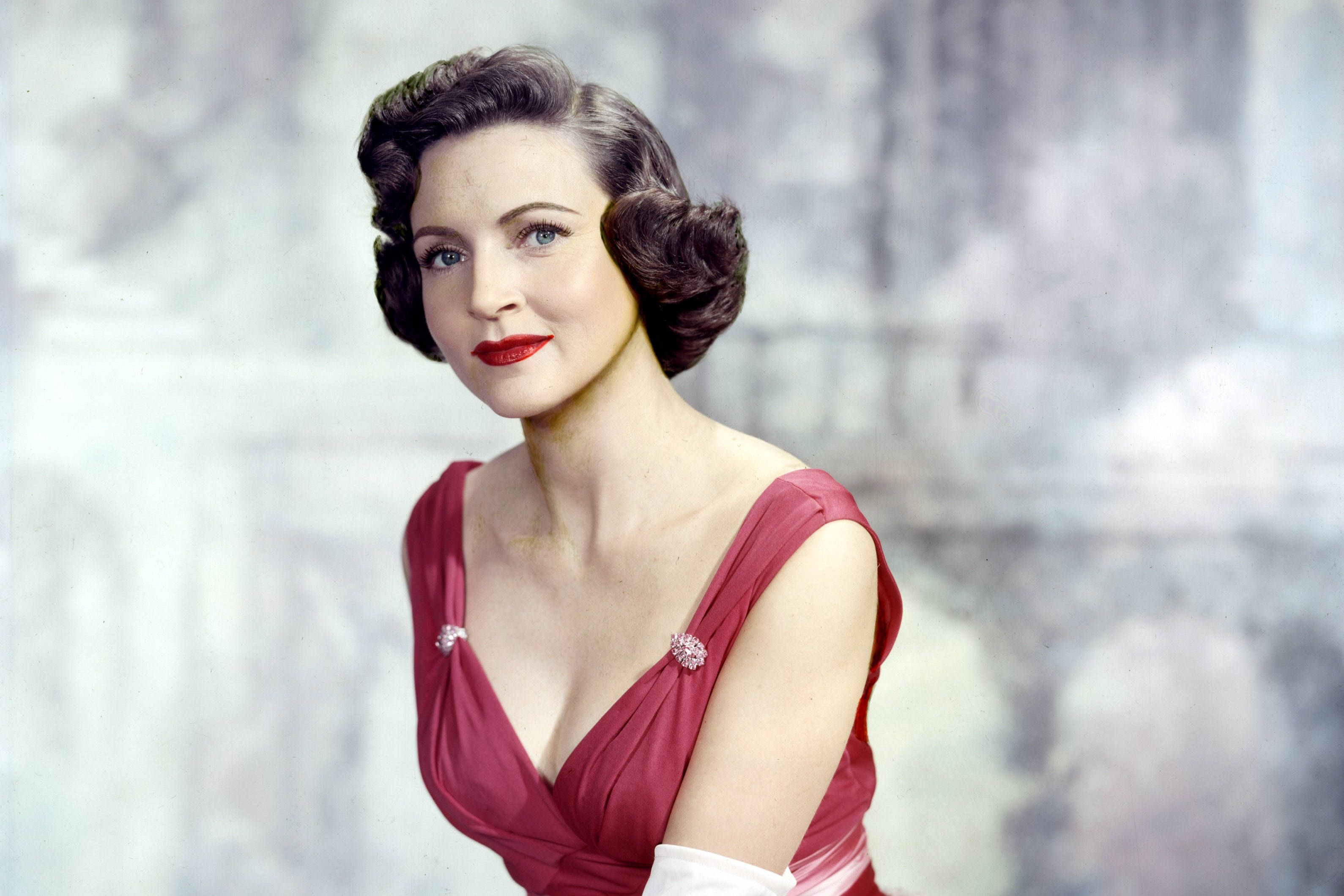 Betty White 桌布 壁紙 — 一位穿著紅色連衣裙、擁有捲曲頭髮和大膽紅唇的魅力女性，在柔和模糊的背景前優雅地擺姿勢。