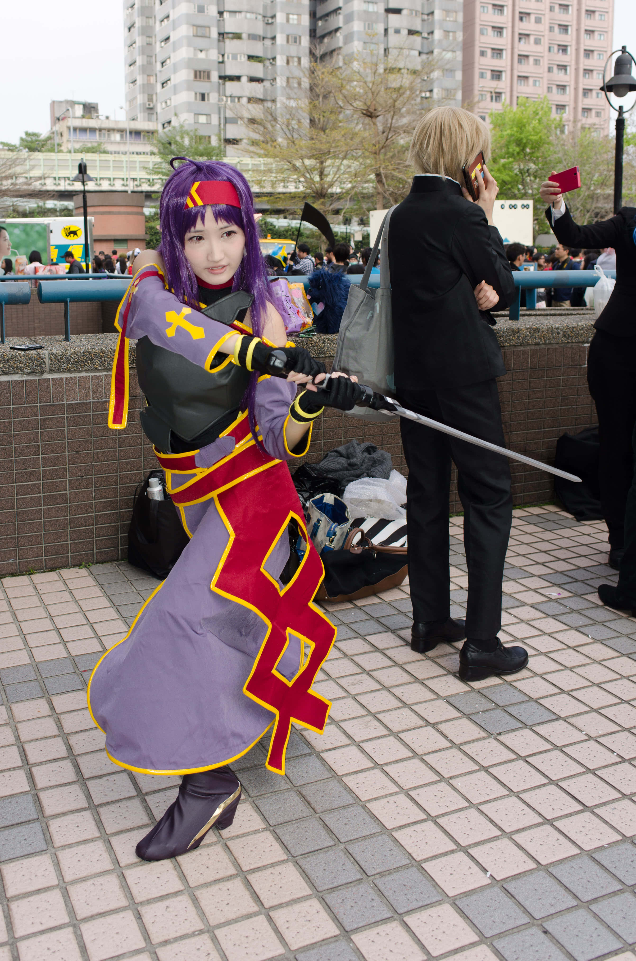 Sword Art Online Cosplay 桌布 壁紙 — 一位穿著紫紅金色奇幻服裝的角色扮演者在城市廣場上揮舞著劍，背後是高樓大廈。