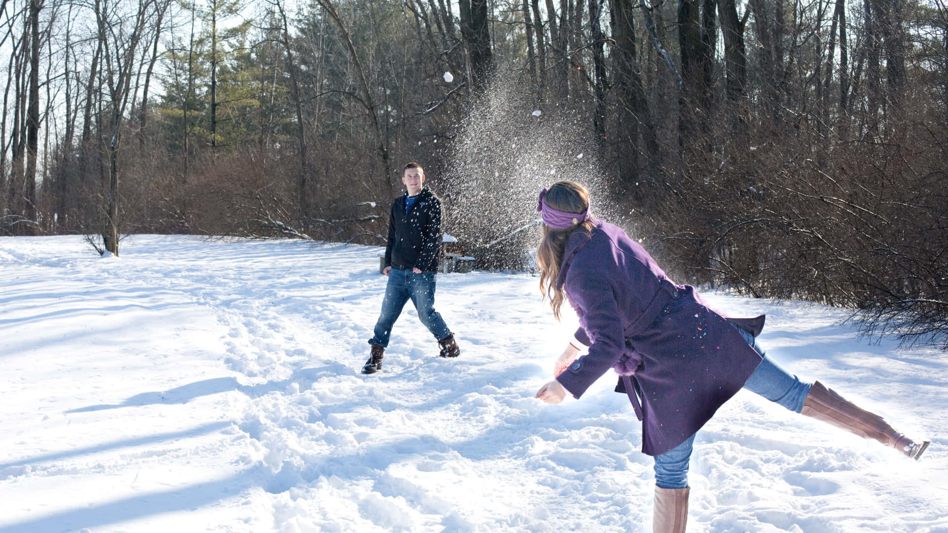 Snowball Fight 桌布 壁紙 — 一位穿著紫色外套的女性在明亮的冬日裡，在白雪覆蓋的森林空地上向一名男子扔雪。