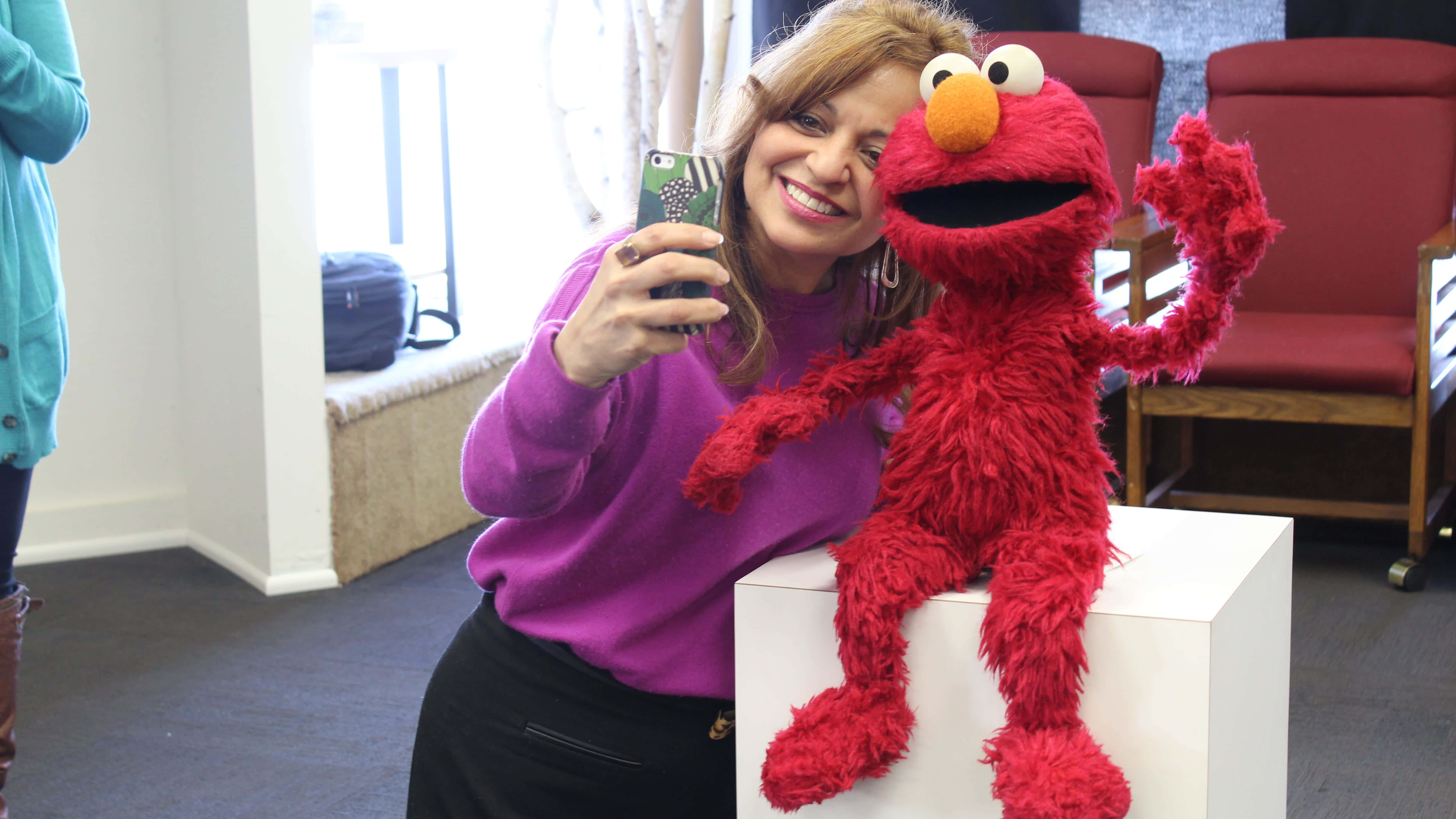 Elmo 桌布 壁紙 — 一位穿著紫色毛衣的微笑女性，在室內與一個坐在白色底座上的大型紅色Elmo公仔自拍。