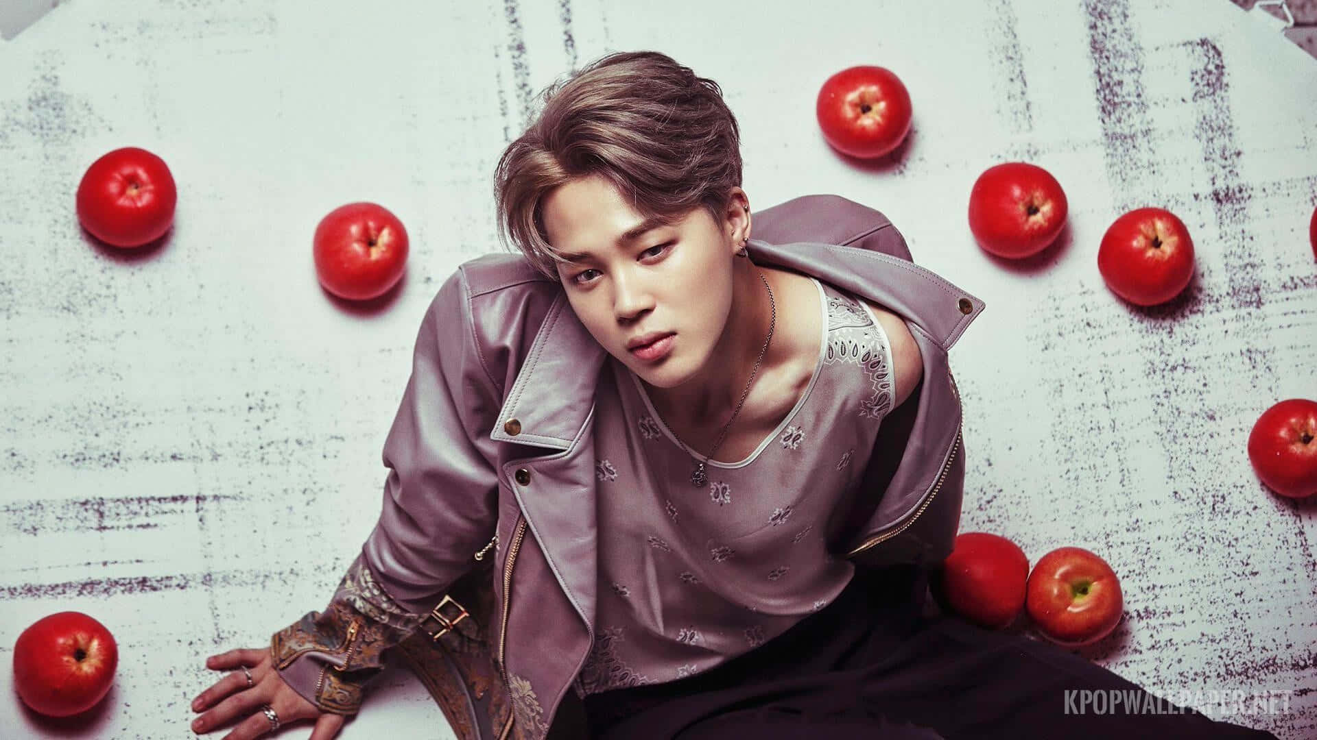 Cute Kpop 桌布 壁紙 — 一位穿著紫色皮夾克的年輕男子在紋理白色地板上被紅蘋果包圍著擺姿勢。