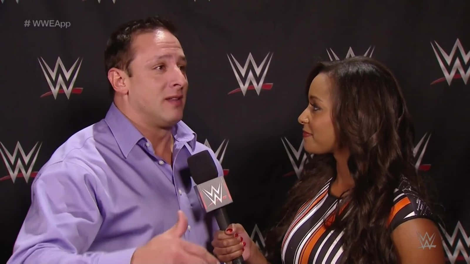 WWE 桌布 壁紙 — 一位穿著紫色襯衫的WWE採訪者對著一位女採訪者手持的麥克風說話，背景為黑色WWE標誌。