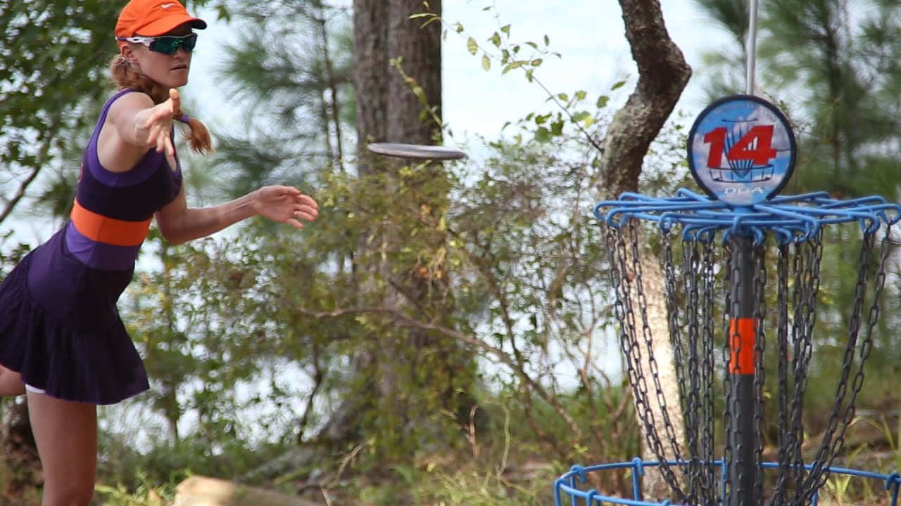 Disc Golf 桌布 壁紙 — 一位穿著紫色連衣裙和橙色帽子的女性，在樹林環繞的戶外環境中，將飛盤擲向一個標有14號的藍色飛盤高爾夫籃框。