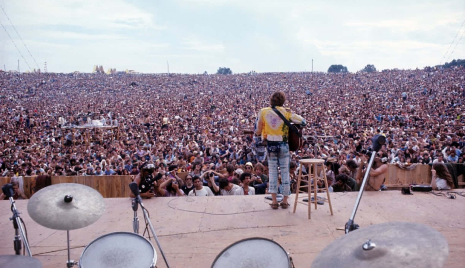 woodstock 桌布 壁紙 — 一位穿著紮染襯衫的音樂家站在舞台上，面對著戶外音樂會中烏雲密布的天空下的大批人群。