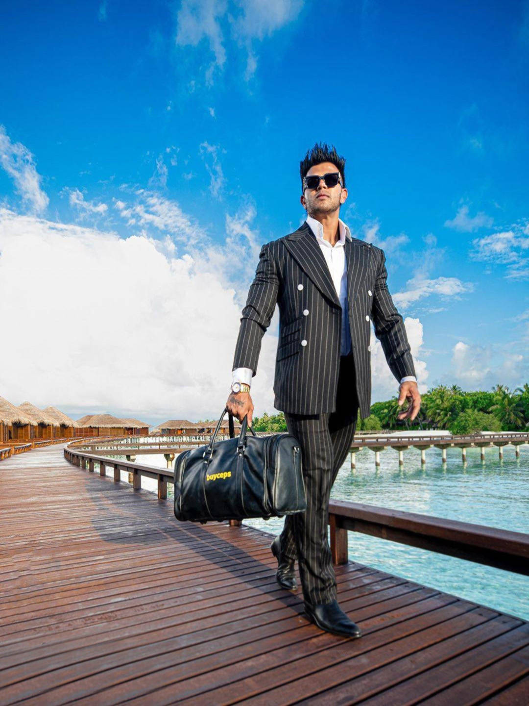 Indian 桌布 壁紙 — 一位穿著細條紋西裝、打扮時尚的男子，在明亮的藍天下，自信地走在碧綠海水上的木質棧道上。