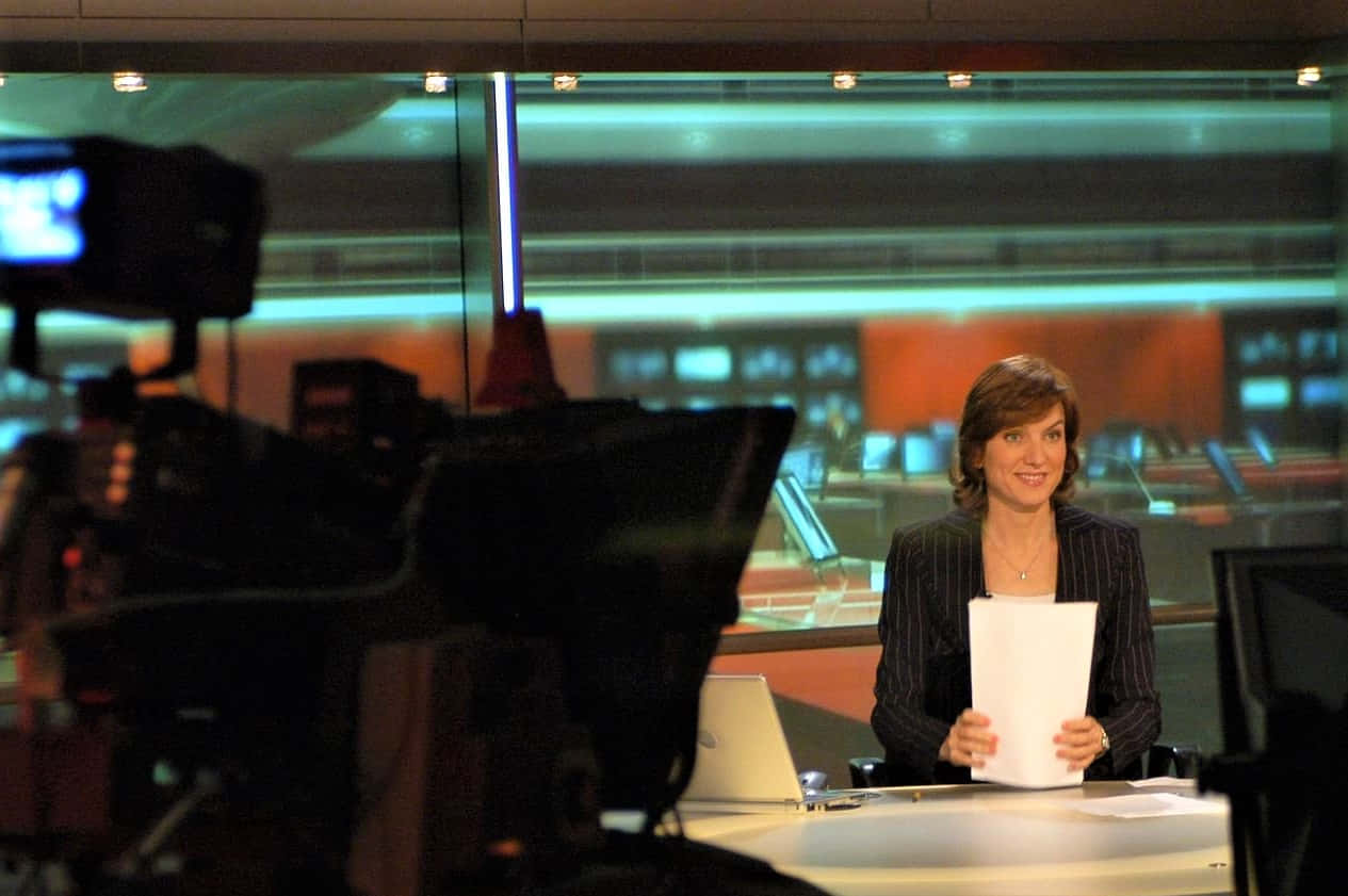 Fiona Bruce 桌布 壁紙 — 一位穿著細條紋西裝、面帶微笑的新聞主播，在現代化的工作室桌前拿著文件，背景有攝影機和發光的綠色燈光。