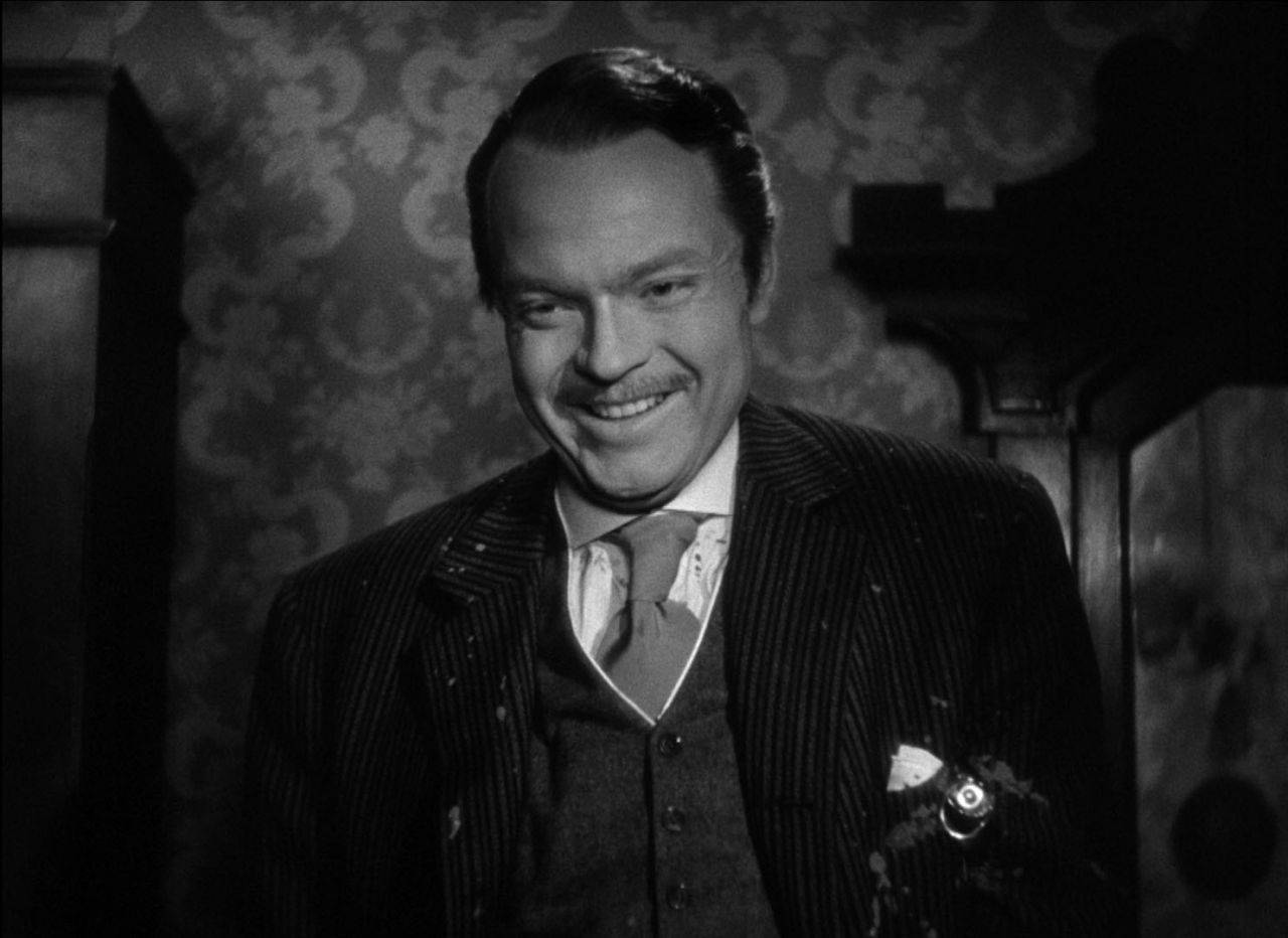 Citizen Kane 桌布 壁紙 — 一位穿著細條紋西裝的男士在光線昏暗的復古房間裡，對著華麗的壁紙溫暖地微笑。