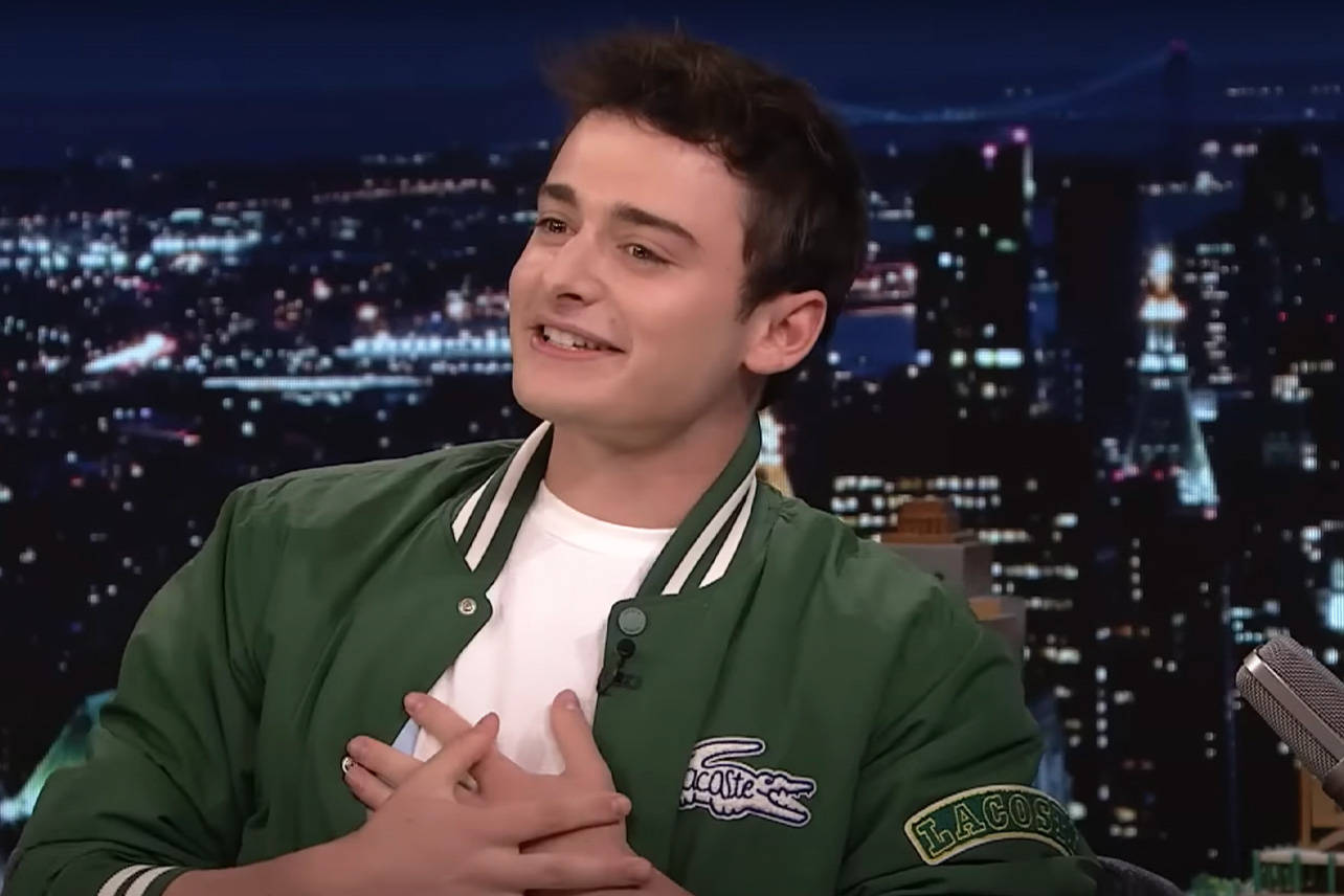 Celebrity 桌布 壁紙 — 一位穿著綠色 Lacoste 外套的年輕男子，坐在充滿活力的夜景城市背景下，露出溫暖的笑容。