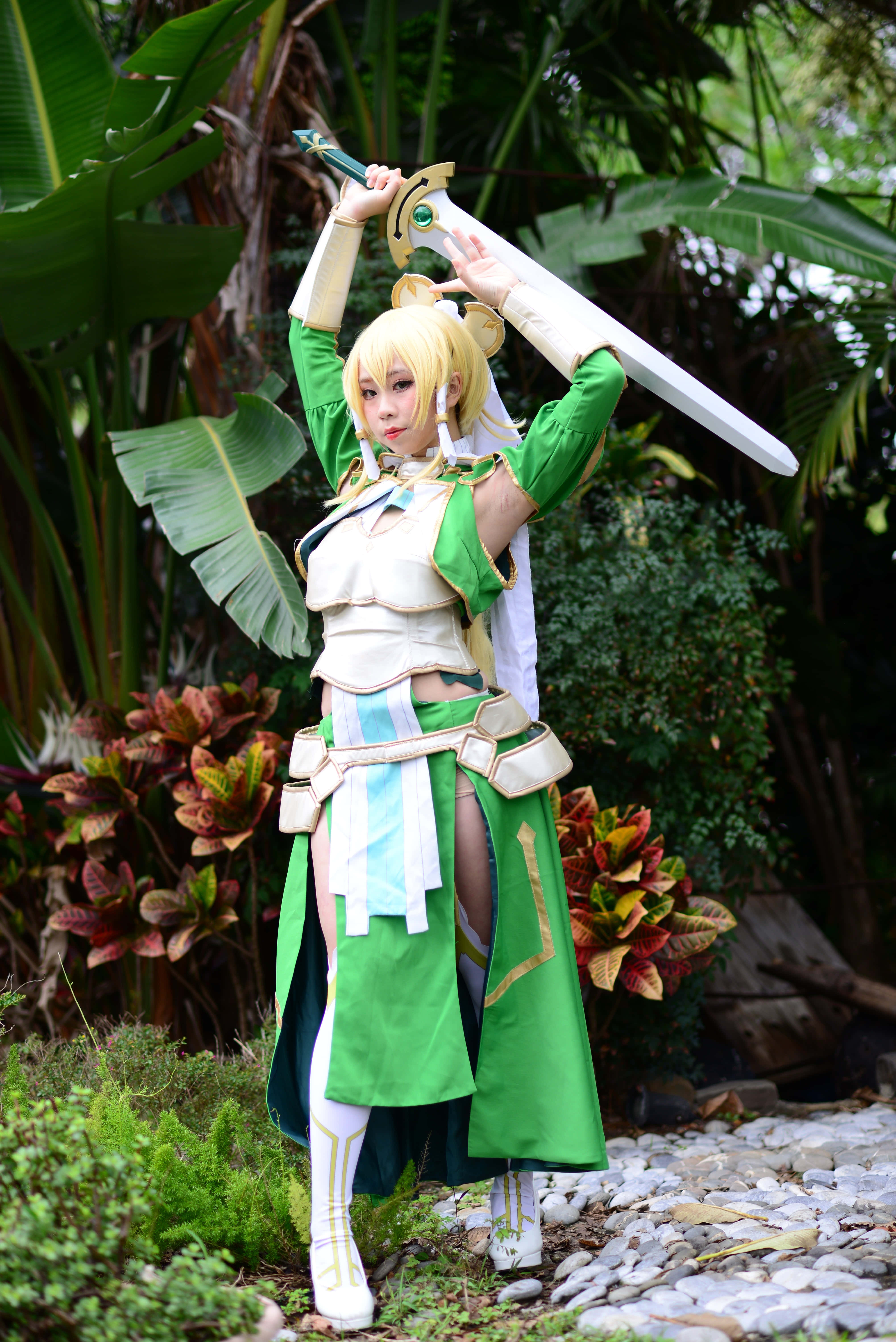 Sword Art Online Cosplay 桌布 壁紙 — 一位穿著綠色和白色奇幻戰士服裝的角色扮演者在茂密的熱帶樹葉中高舉一把大劍。