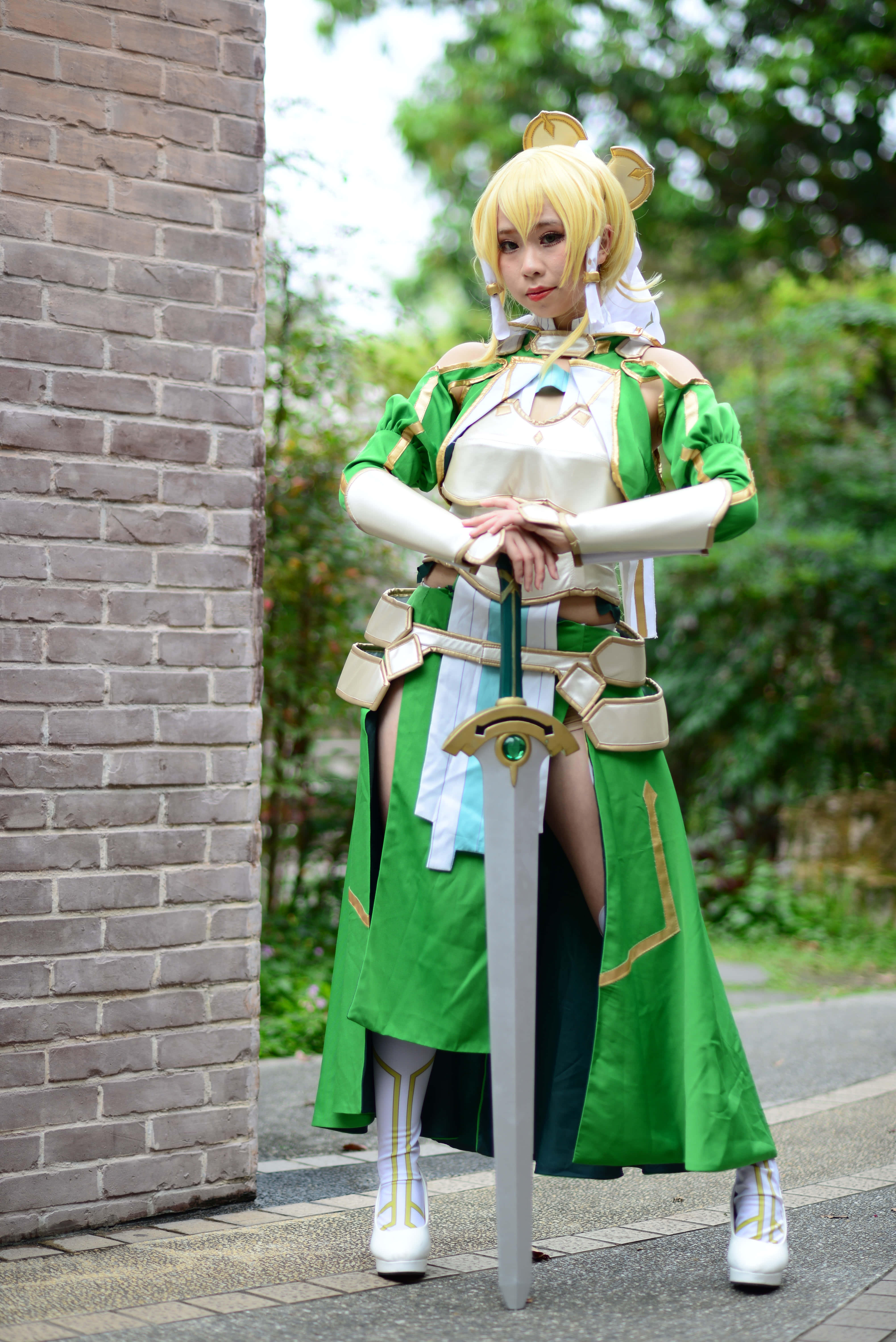 Sword Art Online Cosplay 桌布 壁紙 — 一位穿著綠色和白色奇幻戰士服裝的角色扮演者自信地站在磚牆旁，手持劍。