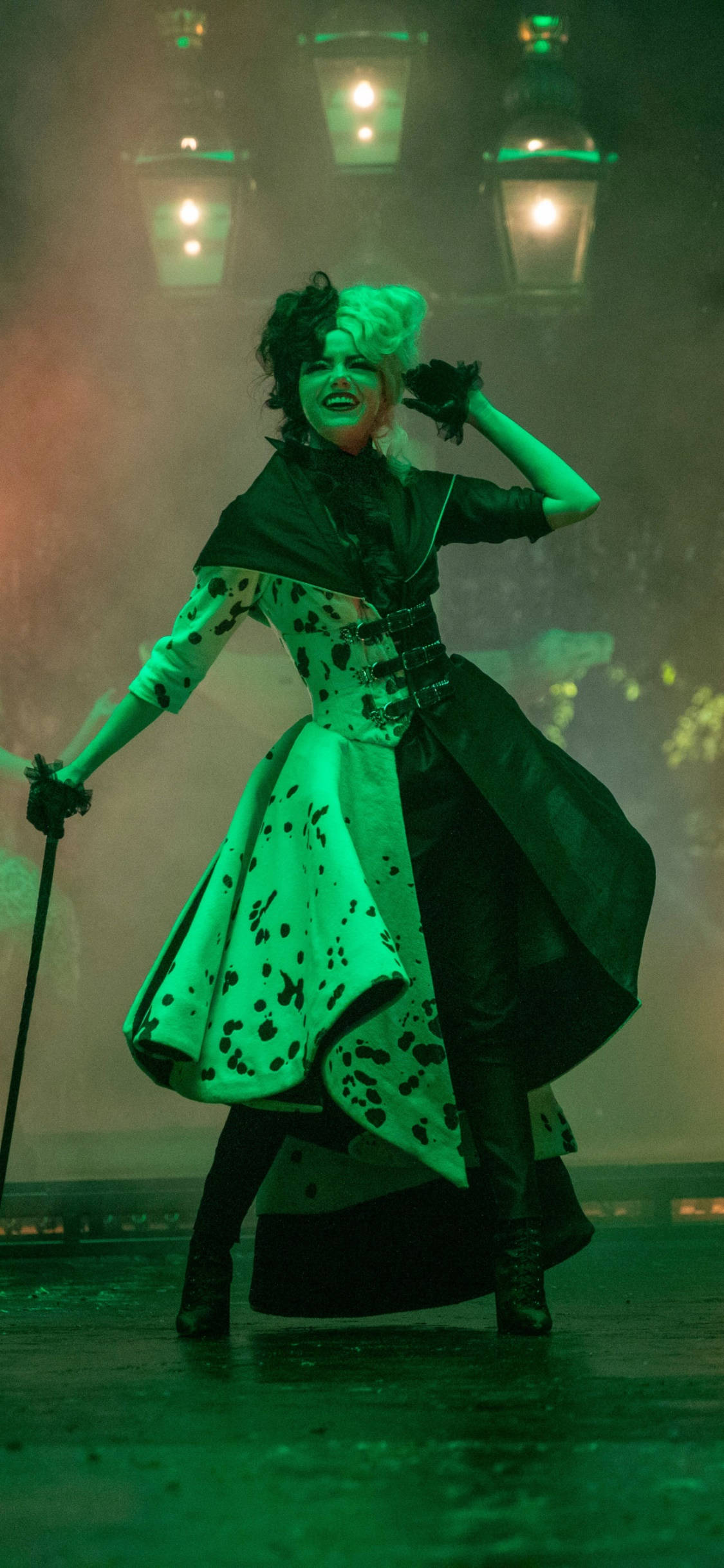 Cruella 2021 桌布 壁紙 — 一位穿著綠色和黑色圓點服裝的表演者在霧氣瀰漫的舞台場景中，在發光的燈籠下擺出姿勢。
