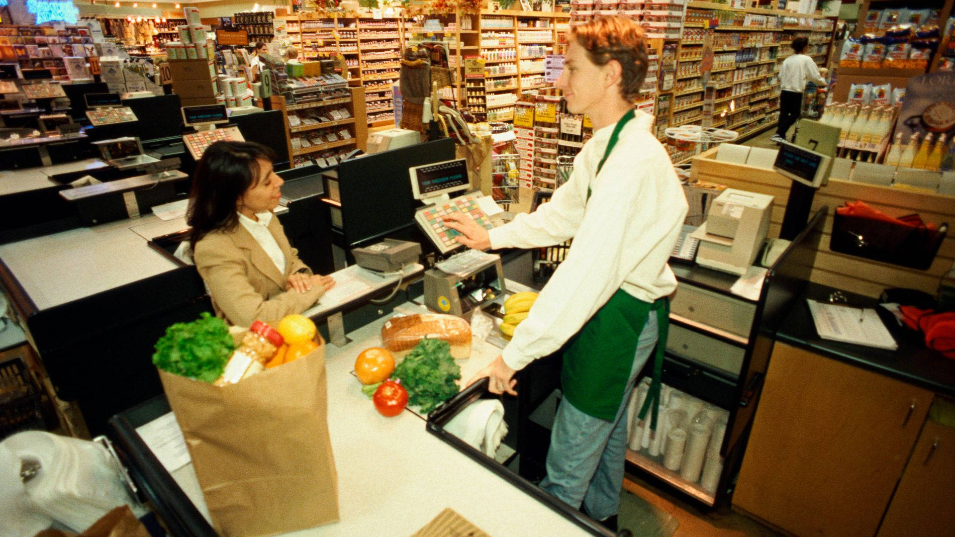 Store 桌布 壁紙 — 一位穿著綠色圍裙的收銀員在超市收銀台掃描雜貨，而顧客耐心等待。