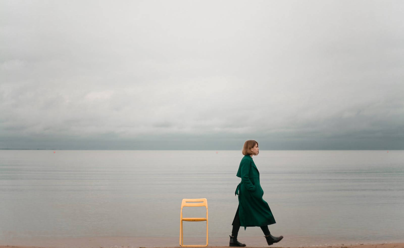 Lonely 桌布 壁紙 — 一位穿著綠色外套的女性在陰天平靜的海灘上走過一張黃色椅子。