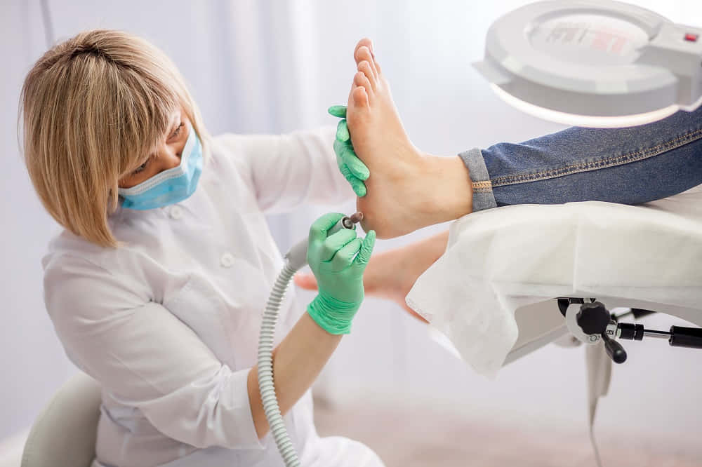 feet 桌布 壁紙 — 一位穿著綠色手套的足病醫生在明亮的臨床環境中使用鑽頭治療病人的腳。