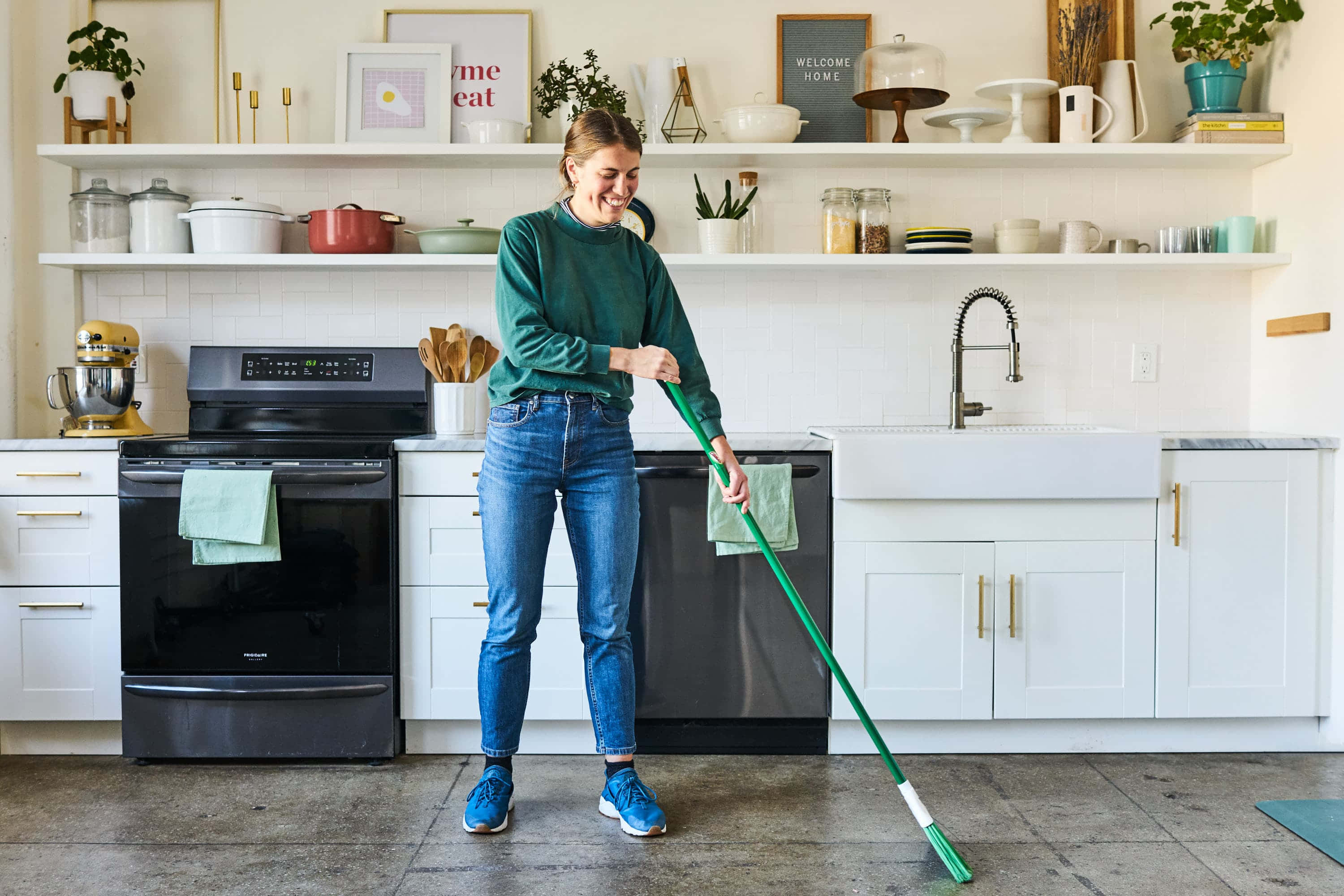 Clean 桌布 壁紙 — 一位穿著綠色毛衣、面帶微笑的女性正在拖地，廚房現代化，配有白色櫥櫃和開放式層架，展示植物和裝飾品。