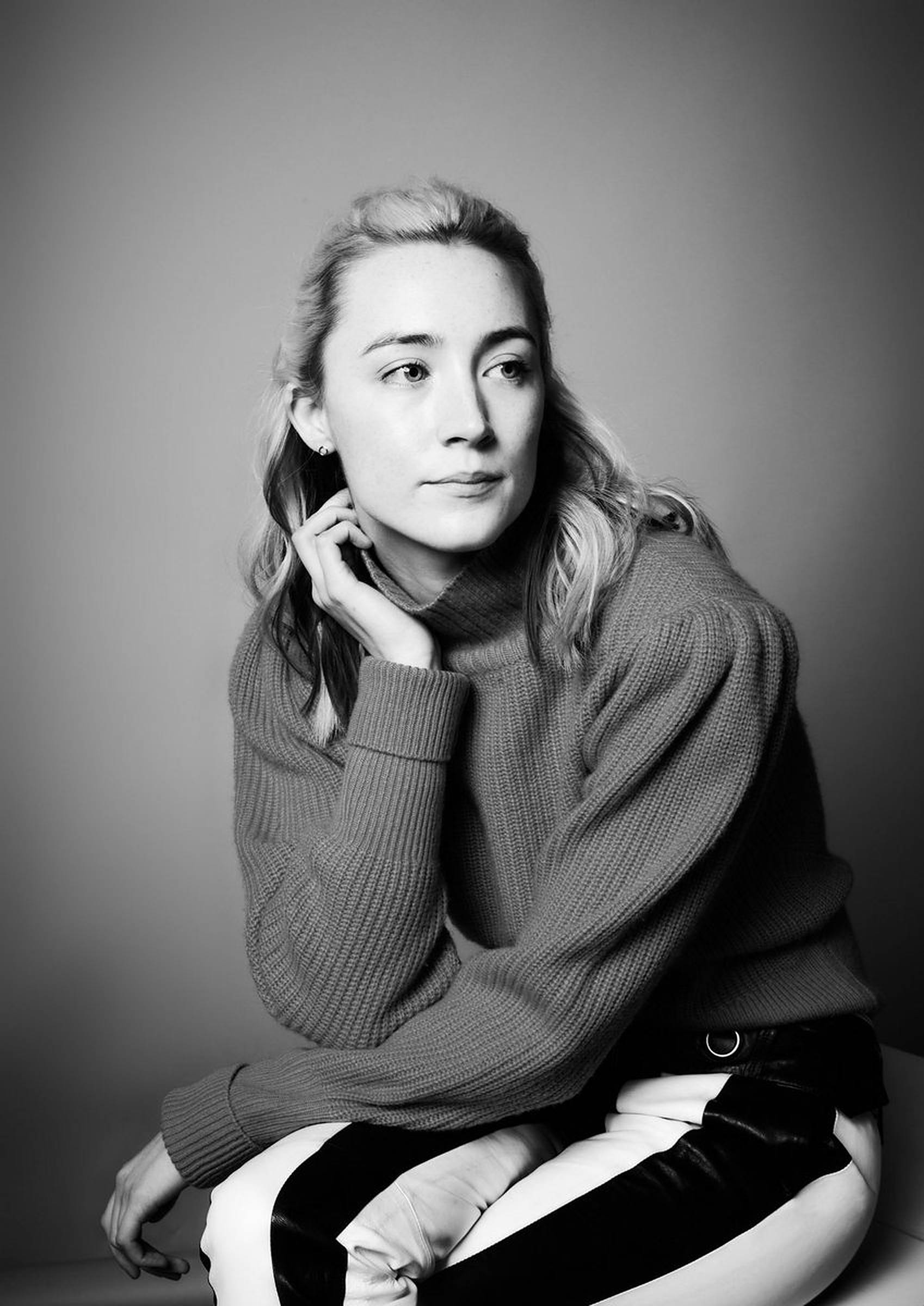 Saoirse Ronan 桌布 壁紙 — 一位穿著羅紋毛衣的女性，在純灰色背景前若有所思地坐著，呈現出引人注目的黑白肖像。