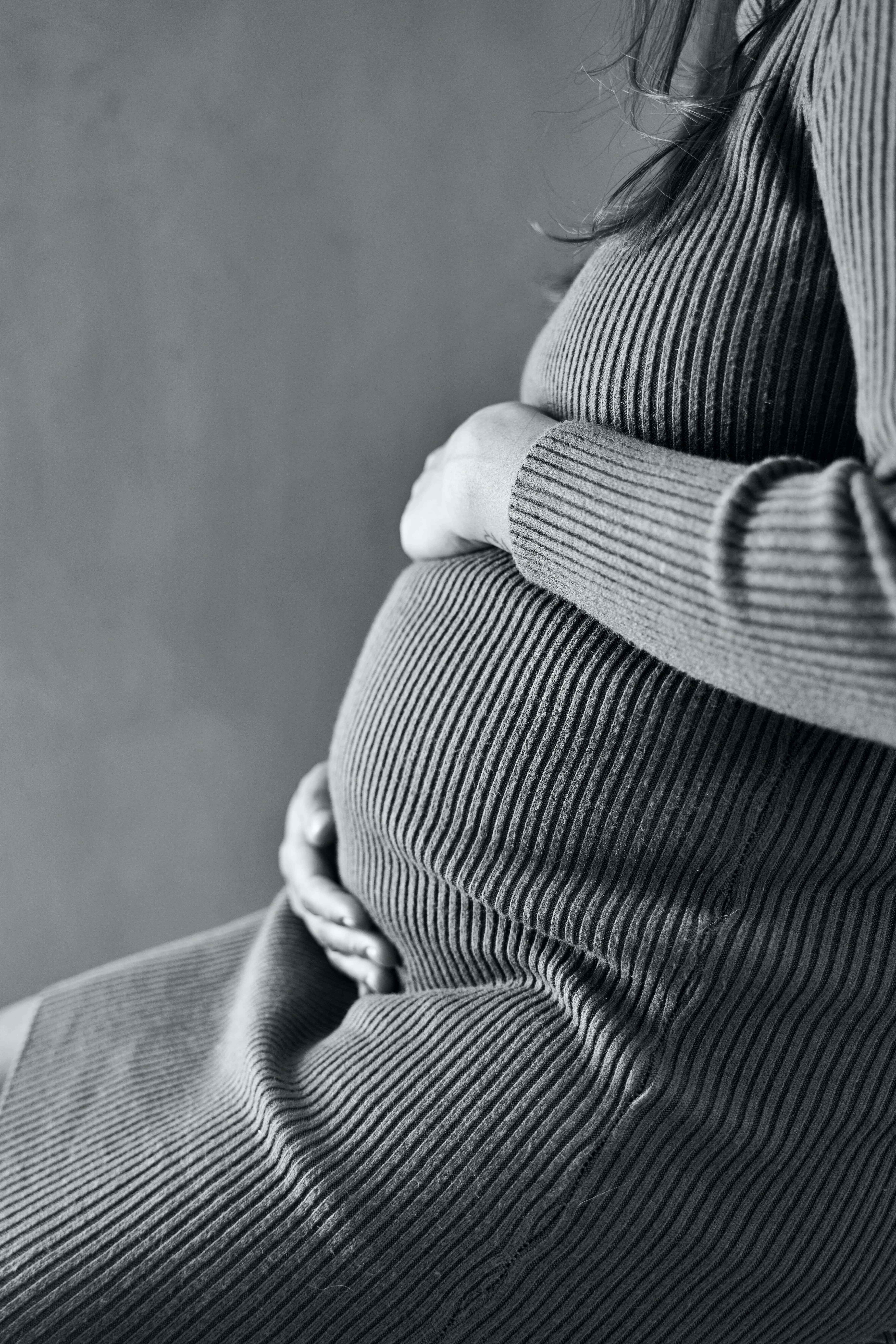 Pregnant 桌布 壁紙 — 一位穿著羅紋毛衣的孕婦，在柔和的灰色背景下，溫柔地抱著肚子，一幅溫柔的黑白肖像。