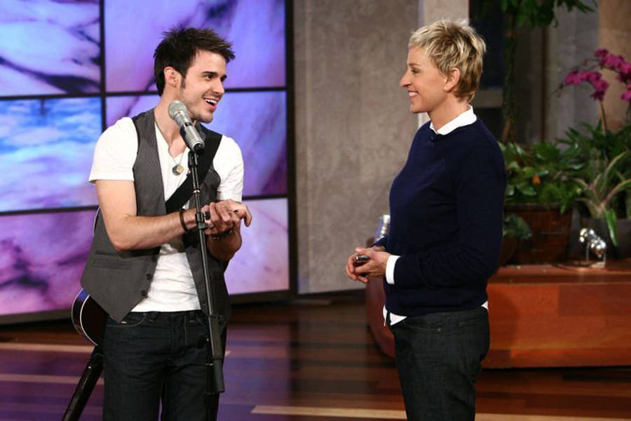 the ellen show 桌布 壁紙 — 一位穿著背心、面帶微笑的男音樂家站在脫口秀佈景中的麥克風旁，與艾倫·狄珍妮交談。