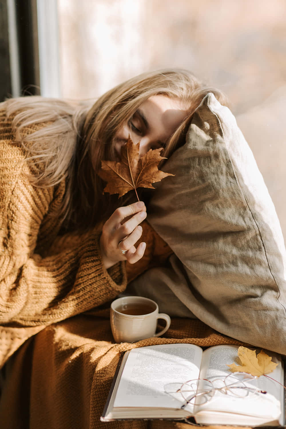 秋季 桌布 壁紙 — 一位穿著舒適毛衣的女性將頭靠在枕頭上，手持一片楓葉靠近臉龐，旁邊放著一本打開的書和咖啡杯。