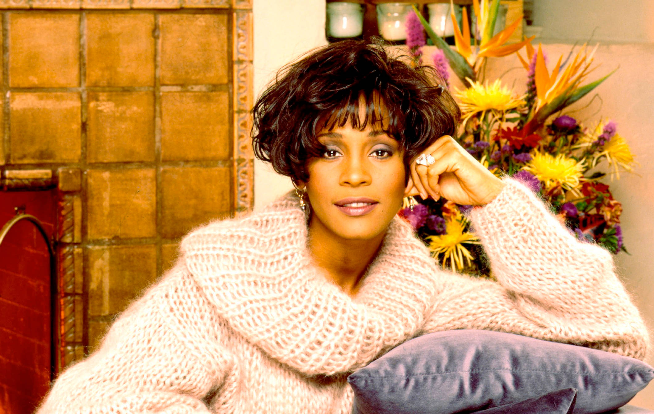 Whitney 桌布 壁紙 — 一位穿著舒適粉紅色針織毛衣的女性，在充滿活力的花卉佈置旁將下巴擱在手上。