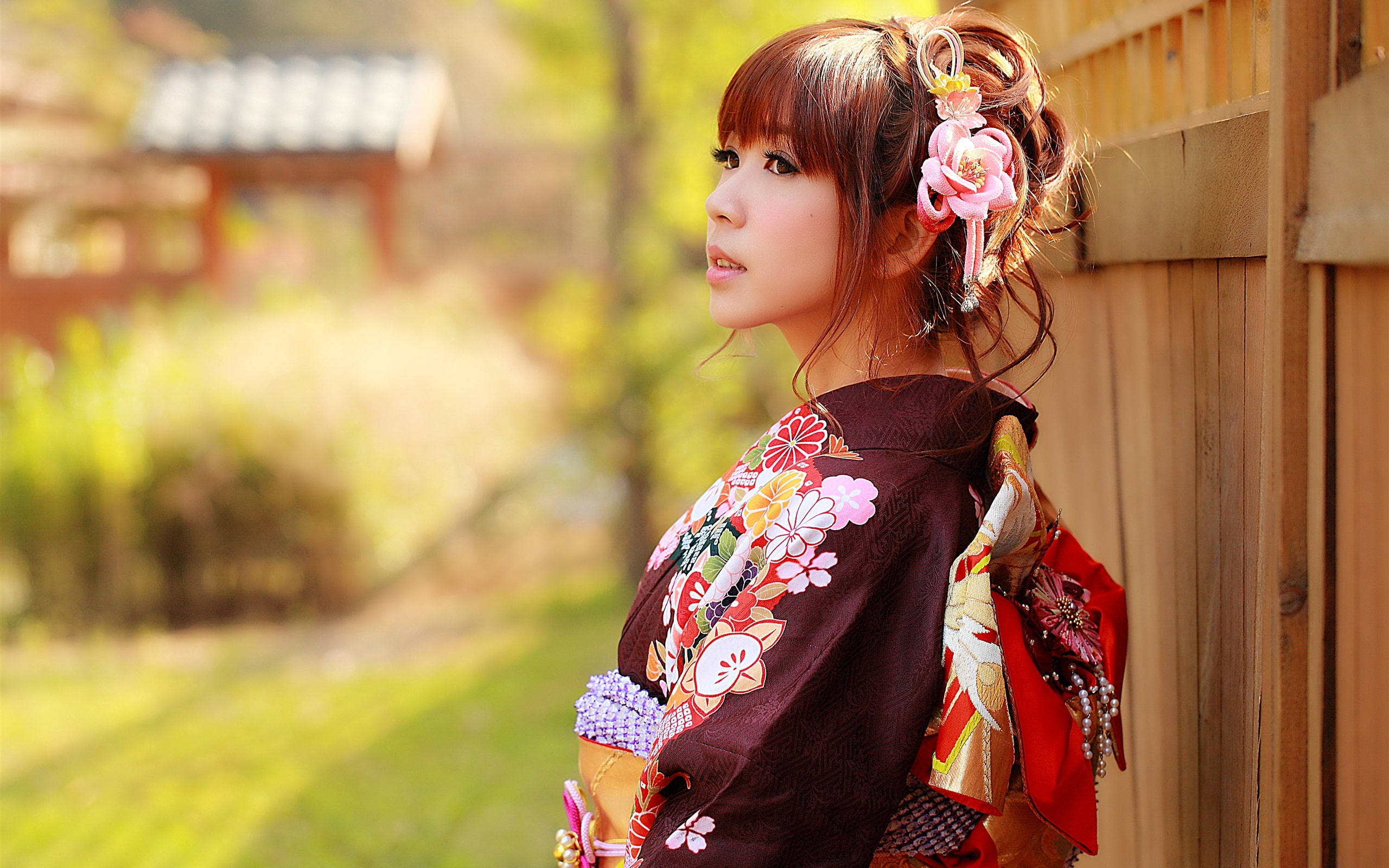 日本 桌布 壁紙 — 一位穿著花卉和服的年輕女子在陽光明媚的花園中靠著木柵欄，背景柔和散景。