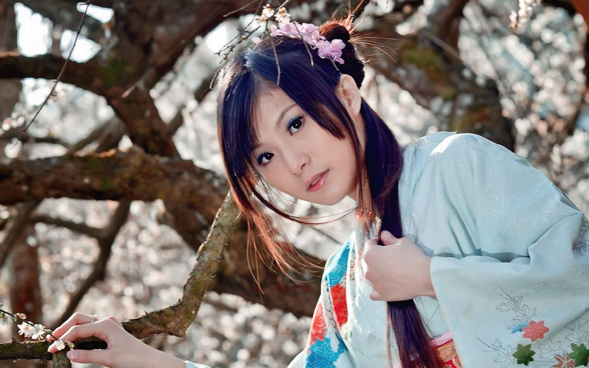 Cute Girl 桌布 壁紙 — 一位穿著花卉和服的年輕女性在柔和陽光下輕輕觸摸樹枝上的櫻花。