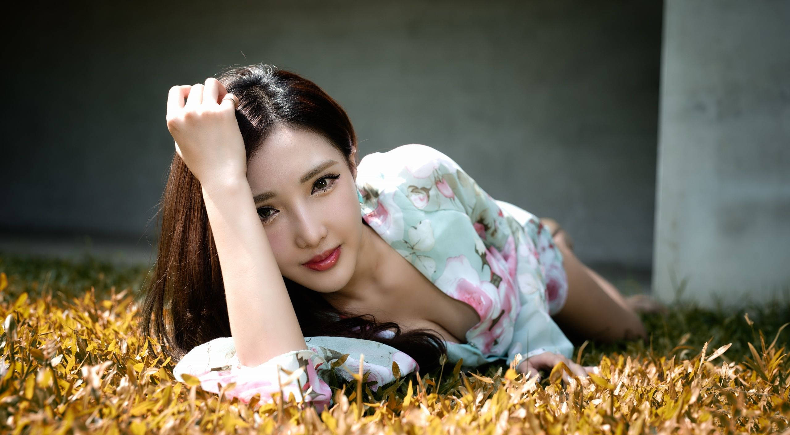 美女 桌布 壁紙 — 一位穿著花卉連衣裙的女性躺在金色草地上，一隻手放在頭上，溫柔地凝視著相機。