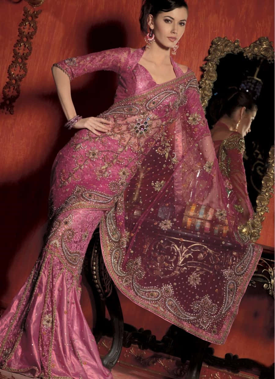 Saree 桌布 壁紙 — 一位穿著華麗刺繡粉紅色和金色紗麗的女性，在豐富的紅色背景下，優雅地站在華麗的鏡子旁邊。