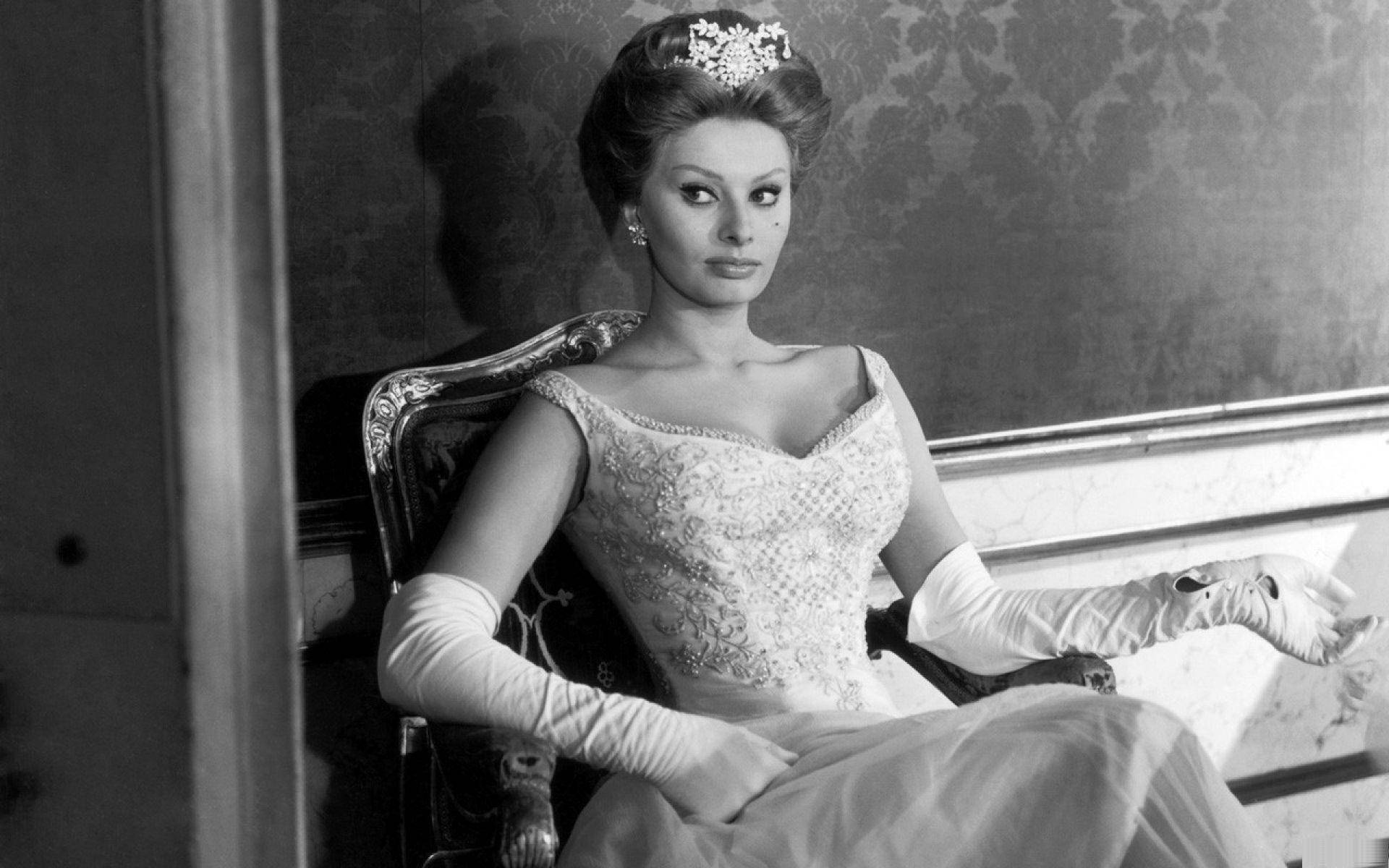 Sophia Loren 桌布 壁紙 — 一位穿著華麗禮服和頭飾的迷人女士，端莊地坐在復古椅子上，背景是圖案壁紙。
