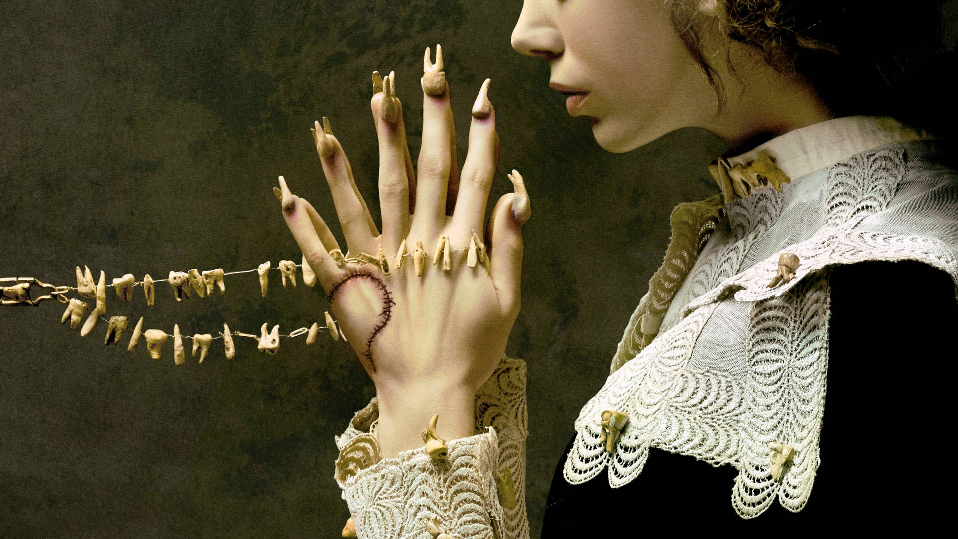 American Horror Story 桌布 壁紙 — 一位穿著華麗蕾絲服裝的女人，用她帶有疤痕的手拿著一串牙齒，靠著深色紋理牆壁。