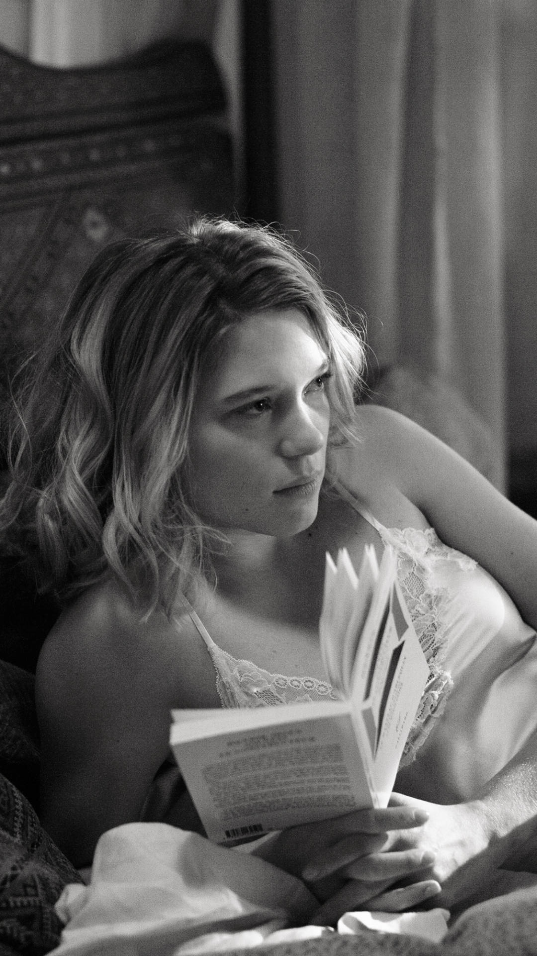 Lea Seydoux 桌布 壁紙 — 一位穿著蕾絲內衣的女性在柔和光線的單色房間裡，若有所思地坐在床上，手裡拿著一本打開的書。