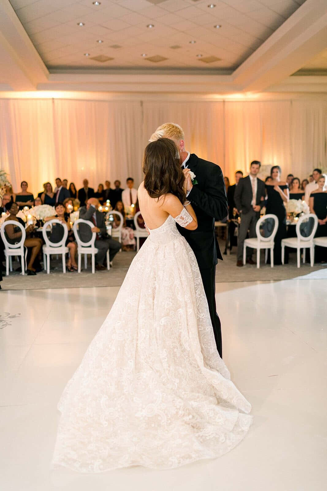 Black And White Wedding 桌布 壁紙 — 一位穿著蕾絲禮服的新娘和一位穿著燕尾服的新郎在白色舞廳地板上分享親密的第一支舞，賓客們在燭光桌旁觀看。