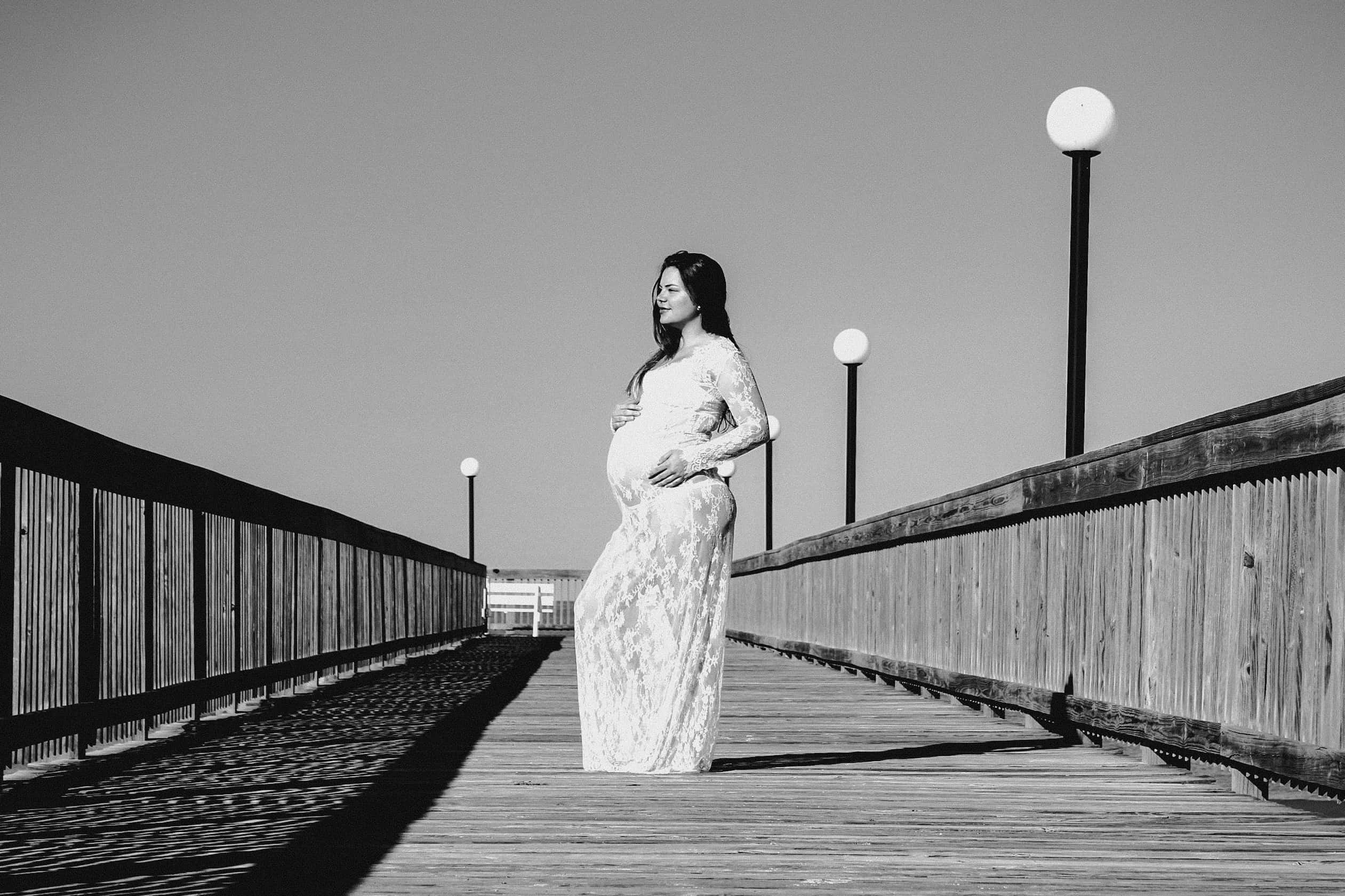 Pregnant 桌布 壁紙 — 一位穿著蕾絲裙的孕婦在晴朗的天空下，優雅地站在一條有街燈排列的木製碼頭上。