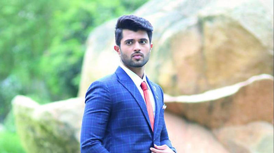vijay devarakonda 4K 桌布 壁紙 — 一位穿著藍色格紋西裝和紅色領帶、打扮俐落的男士，自信地站在大岩石和茂密綠植的背景前。