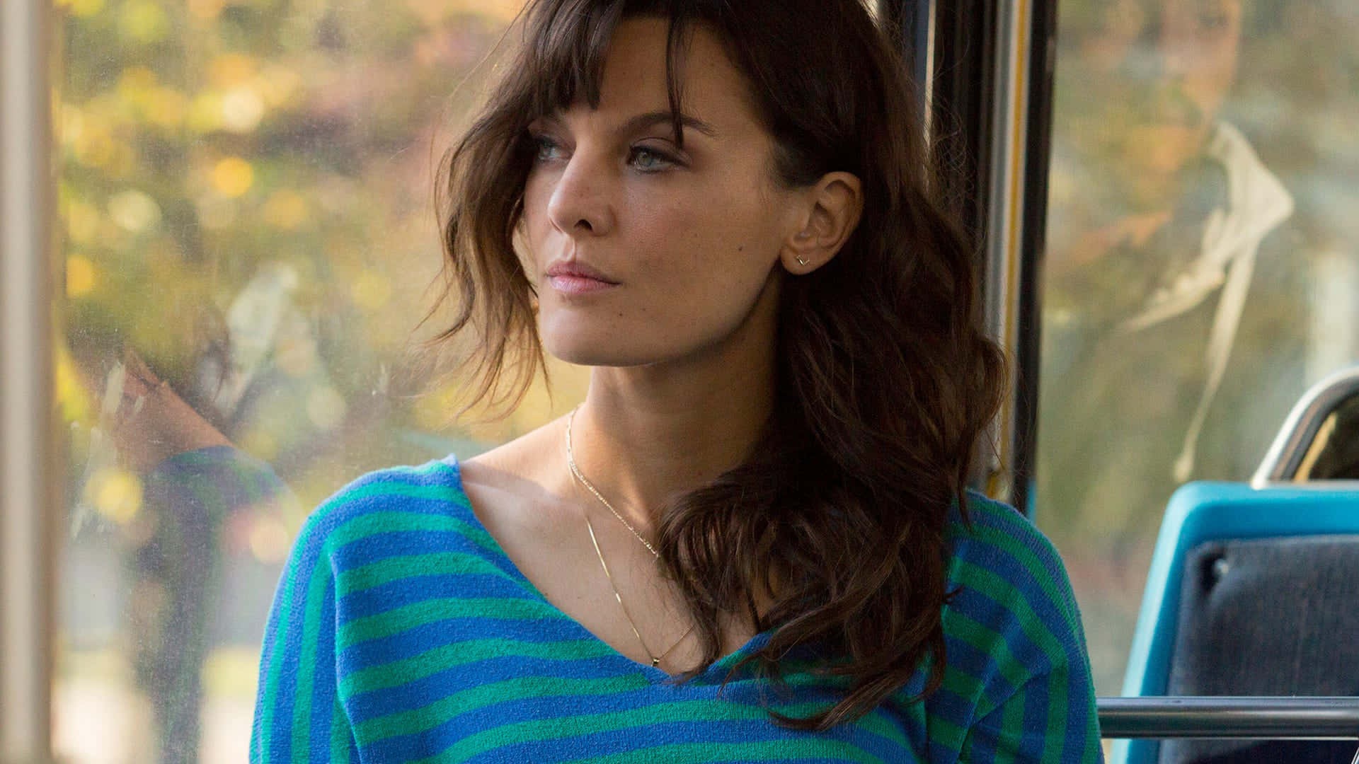 Frankie Shaw 桌布 壁紙 — 一位穿著藍色條紋毛衣的女性若有所思地望著巴士窗外，秋天的樹葉模糊地掠過。
