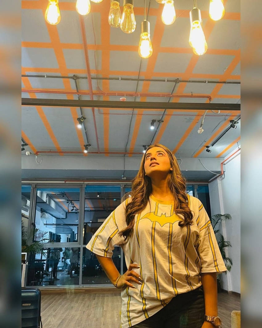 Light Bulb 桌布 壁紙 — 一位穿著蝙蝠俠T恤的女性抬頭望向懸掛在橙色工業風天花板上的發光愛迪生燈泡。