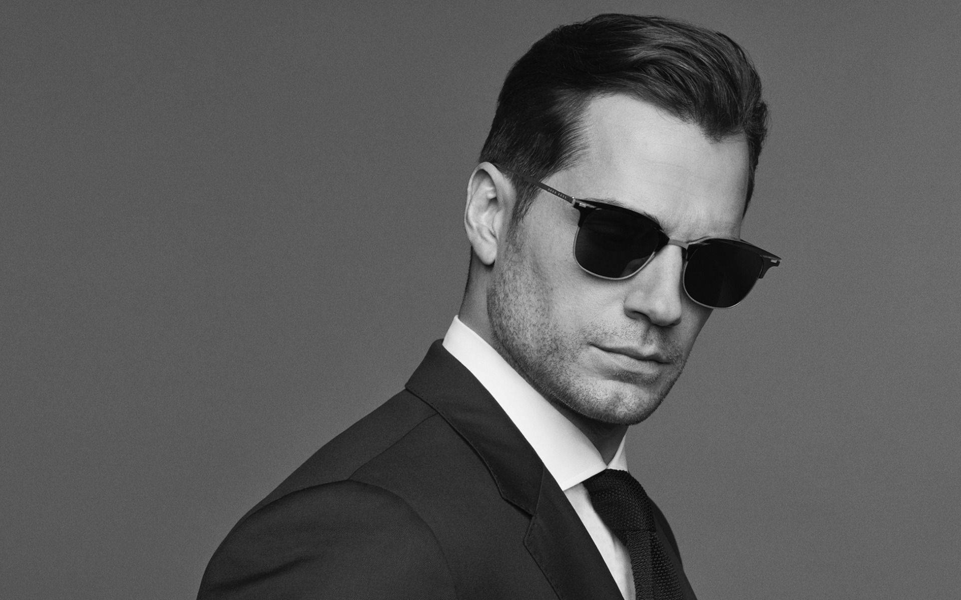 Sunglasses 桌布 壁紙 — 一位穿著西裝和太陽眼鏡、打扮俐落的男士，在純灰色背景前擺出嚴肅的表情。