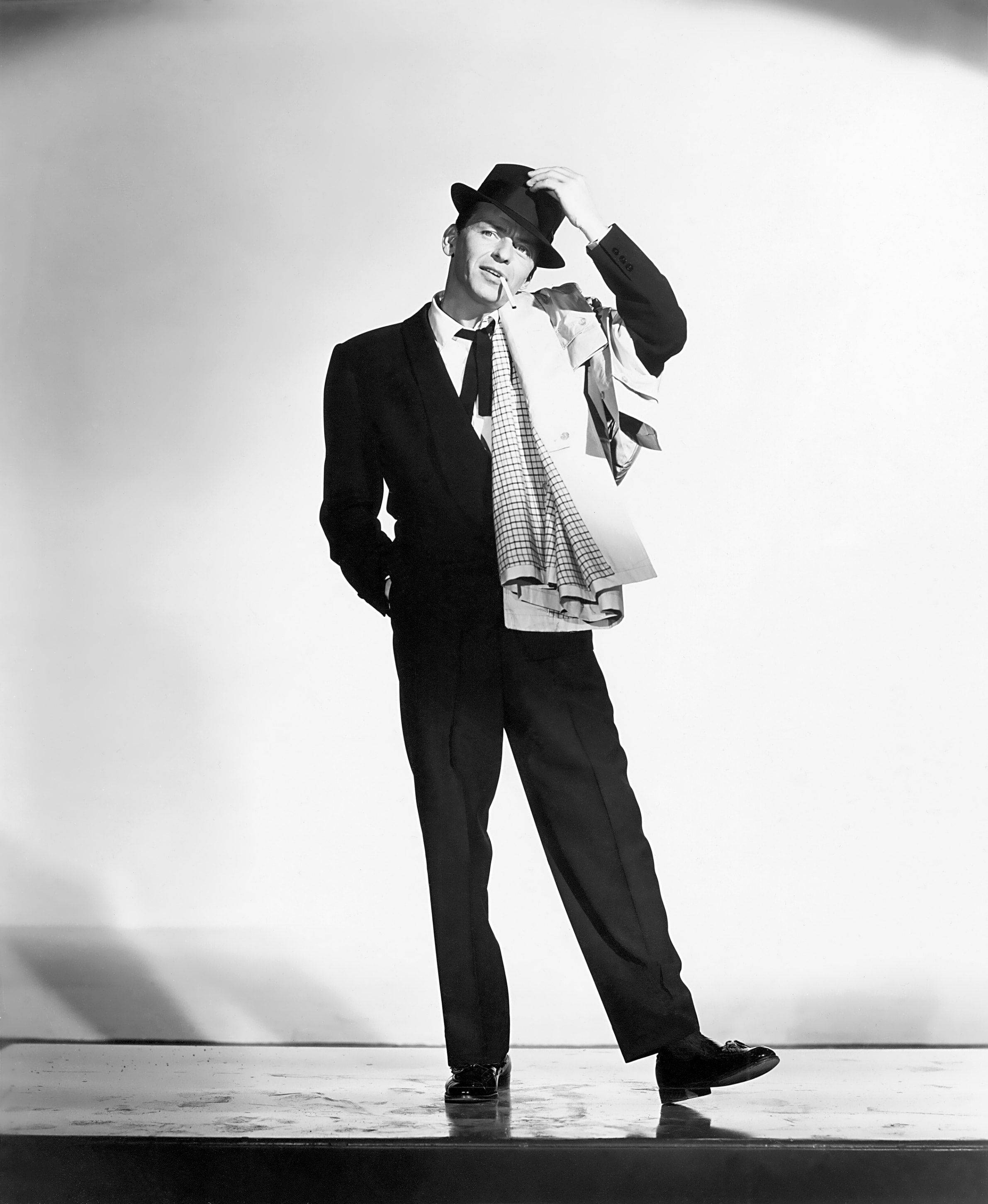 Sinatra 桌布 壁紙 — 一位穿著西裝和軟呢帽的時尚男士，在簡潔的攝影棚背景下自信微笑地傾斜帽子。
