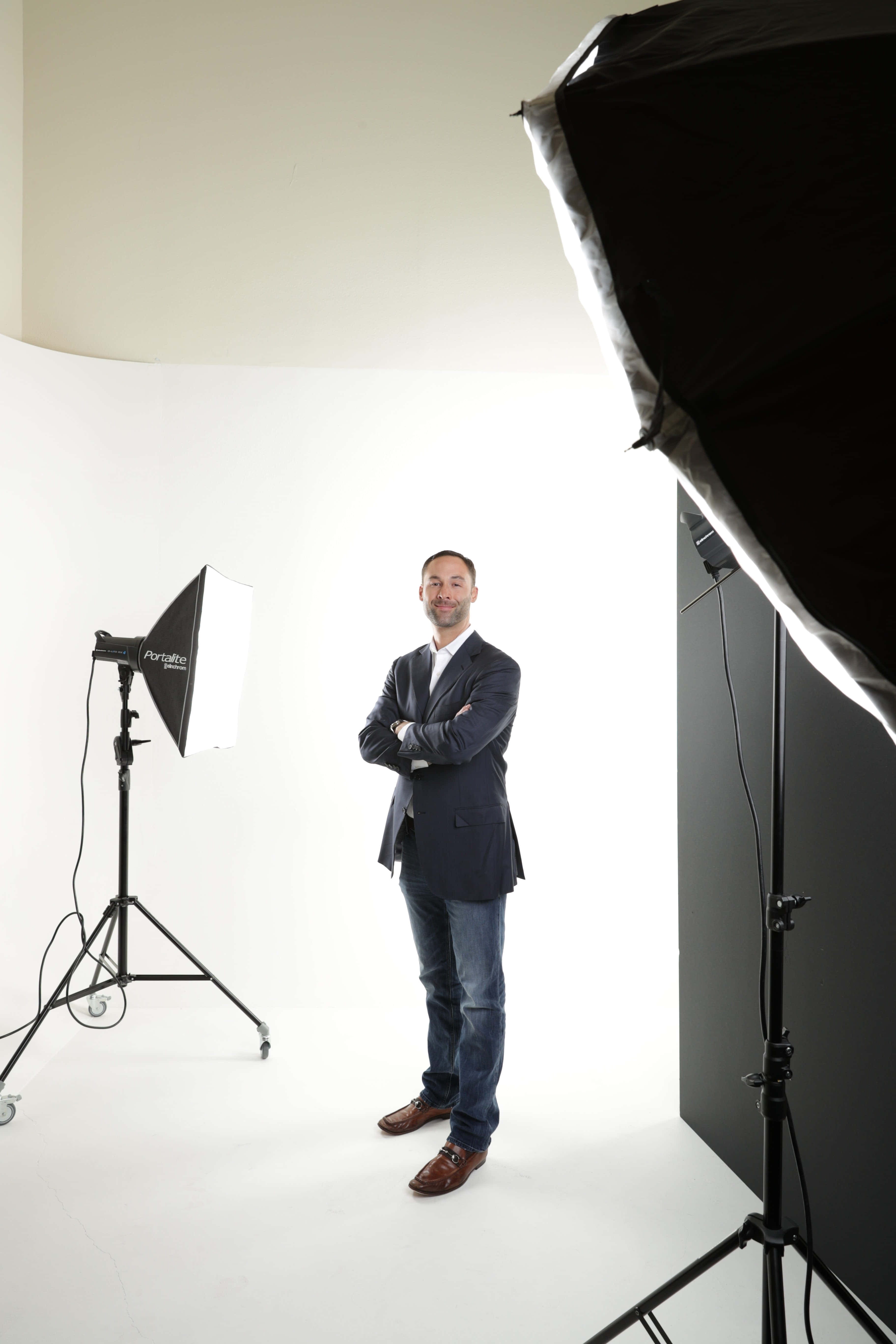 Photography Studio 桌布 壁紙 — 一位穿著西裝外套的男子自信地交叉雙臂，站在明亮的白色攝影棚中，周圍環繞著專業照明設備。