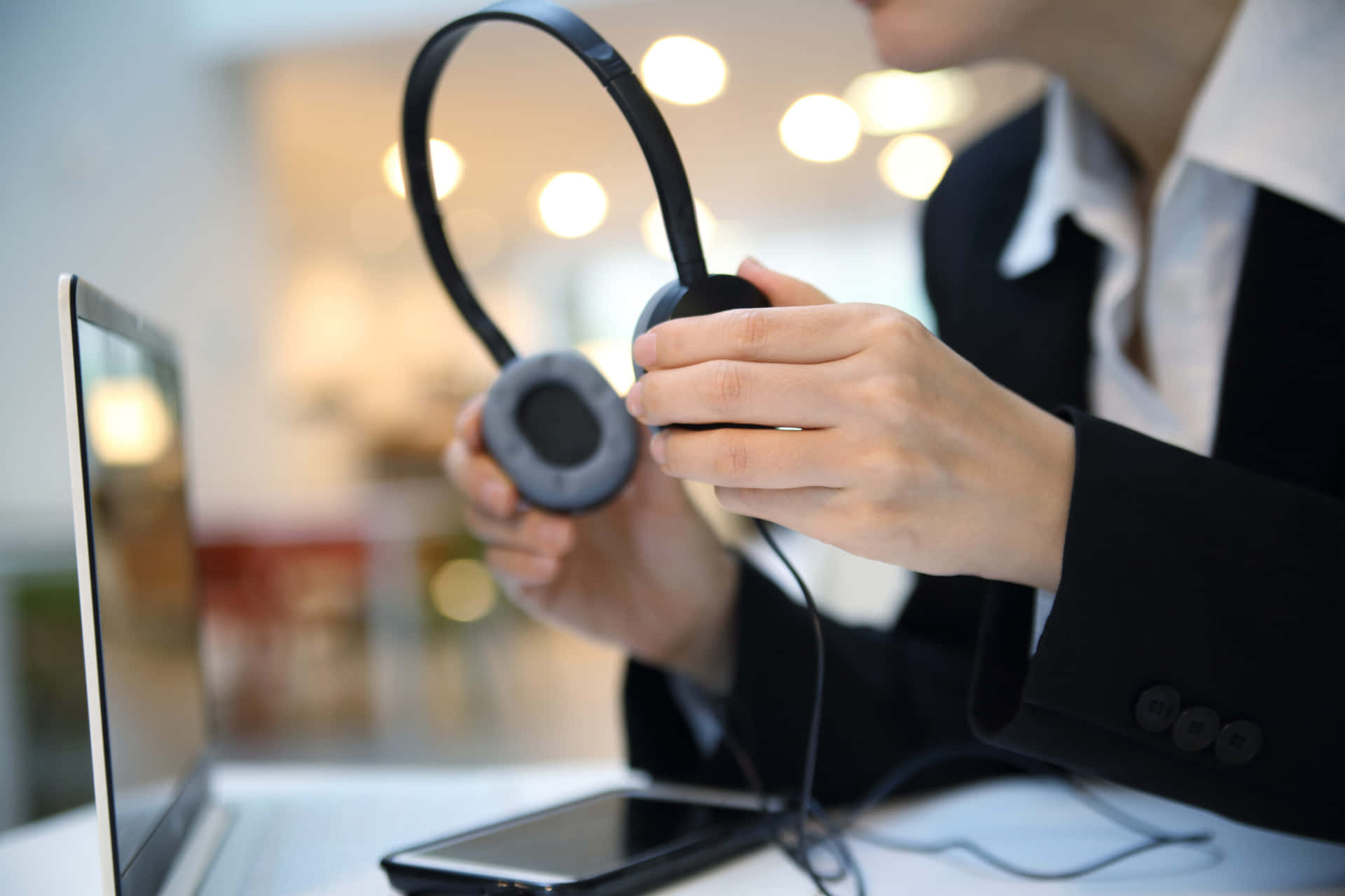 Customer Service 桌布 壁紙 — 一位穿著西裝的人士在柔和光線的現代辦公室中，手持黑色頭戴式耳機靠近筆記型電腦。