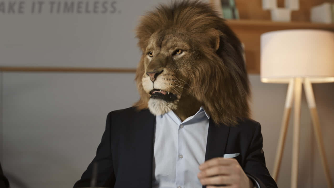 Lion Head 桌布 壁紙 — 一位穿著西裝的人戴著逼真的獅子頭面具，坐在現代辦公室環境中。