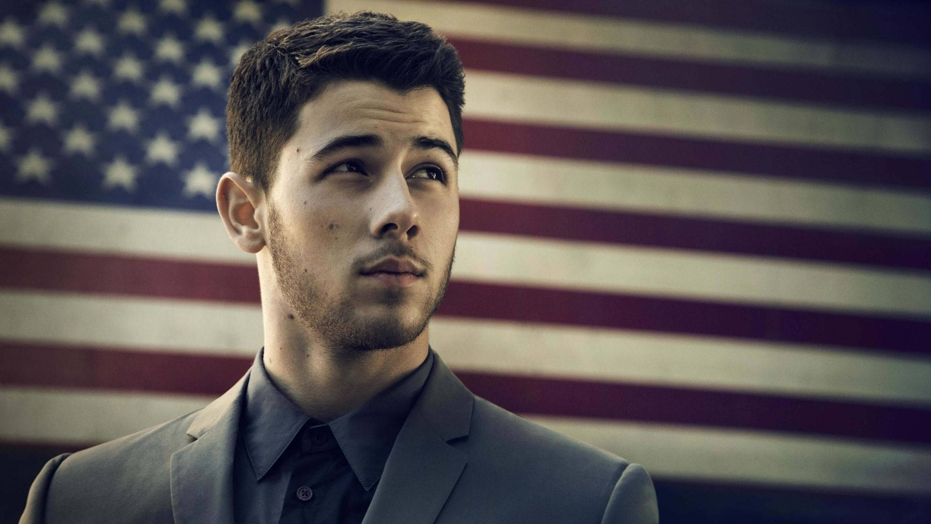 Nick Jonas 桌布 壁紙 — 一位穿著西裝的年輕男子，在模糊的美國國旗背景下若有所思地凝視。
