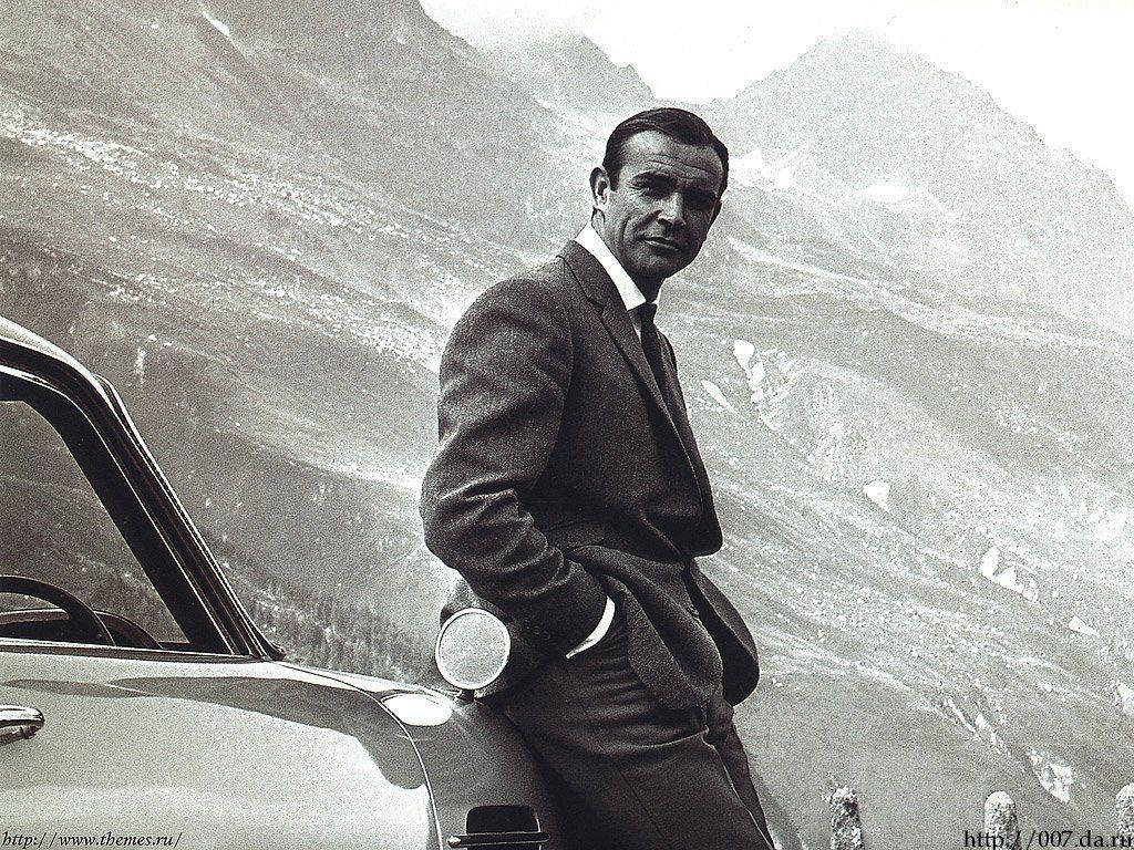Bond 桌布 壁紙 — 一位穿著西裝的時尚男士靠在一輛經典汽車旁，背景是雄偉的山脈，經典黑白照片。