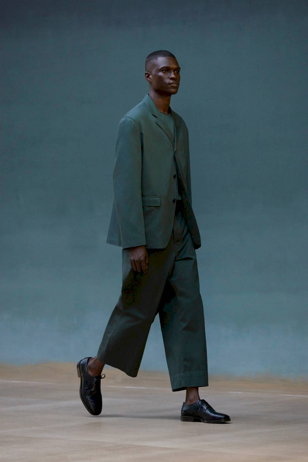 Mens Fashion 桌布 壁紙 — 一位穿著訂製鼠尾草綠色西裝的優雅男士，在柔和青綠色的背景下，自信地走過極簡風格的攝影棚地板。