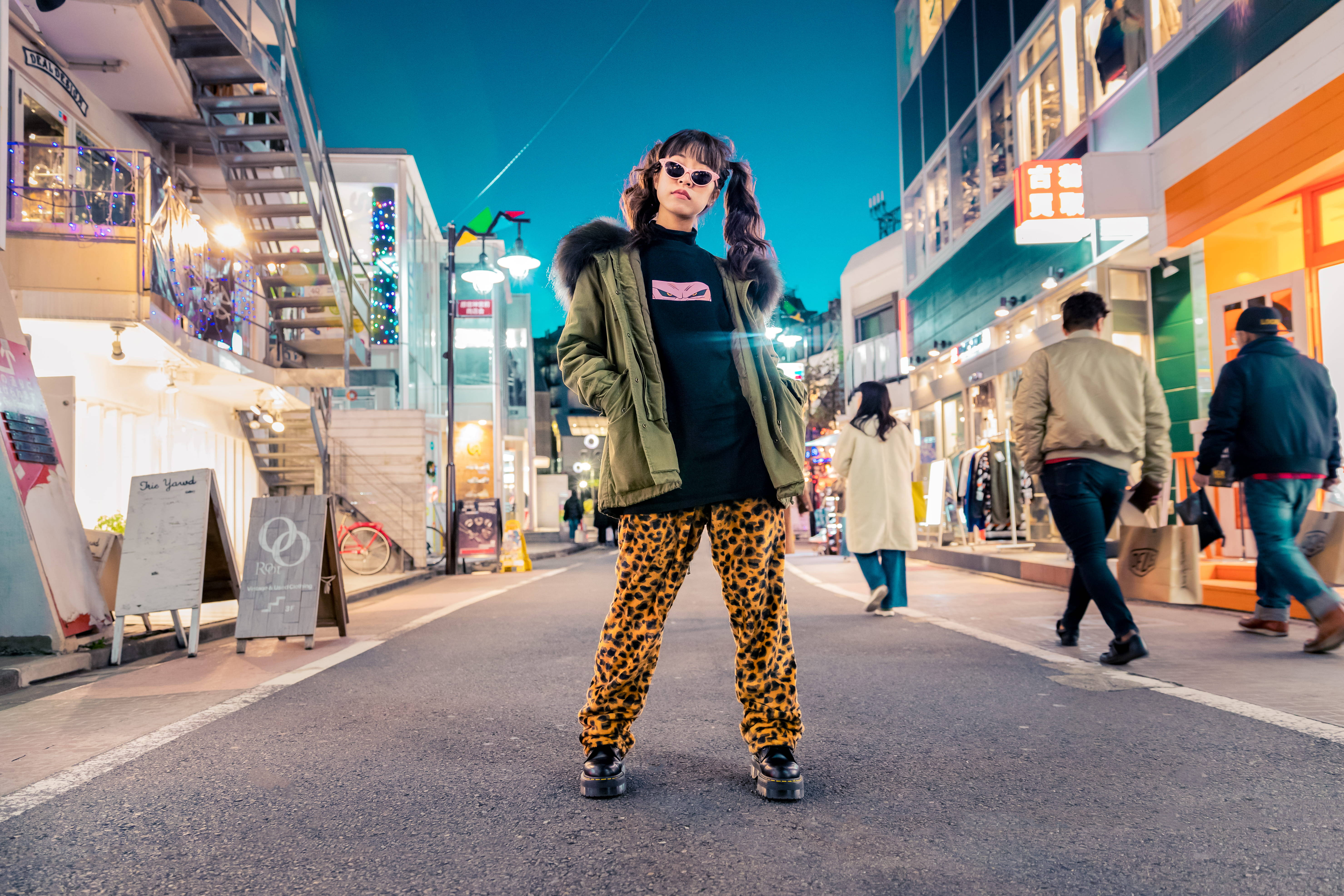 東京 桌布 壁紙 — 一位穿著豹紋褲和綠色外套的時尚人士，在黃昏時分自信地站在霓虹燈閃爍的都市街道上。
